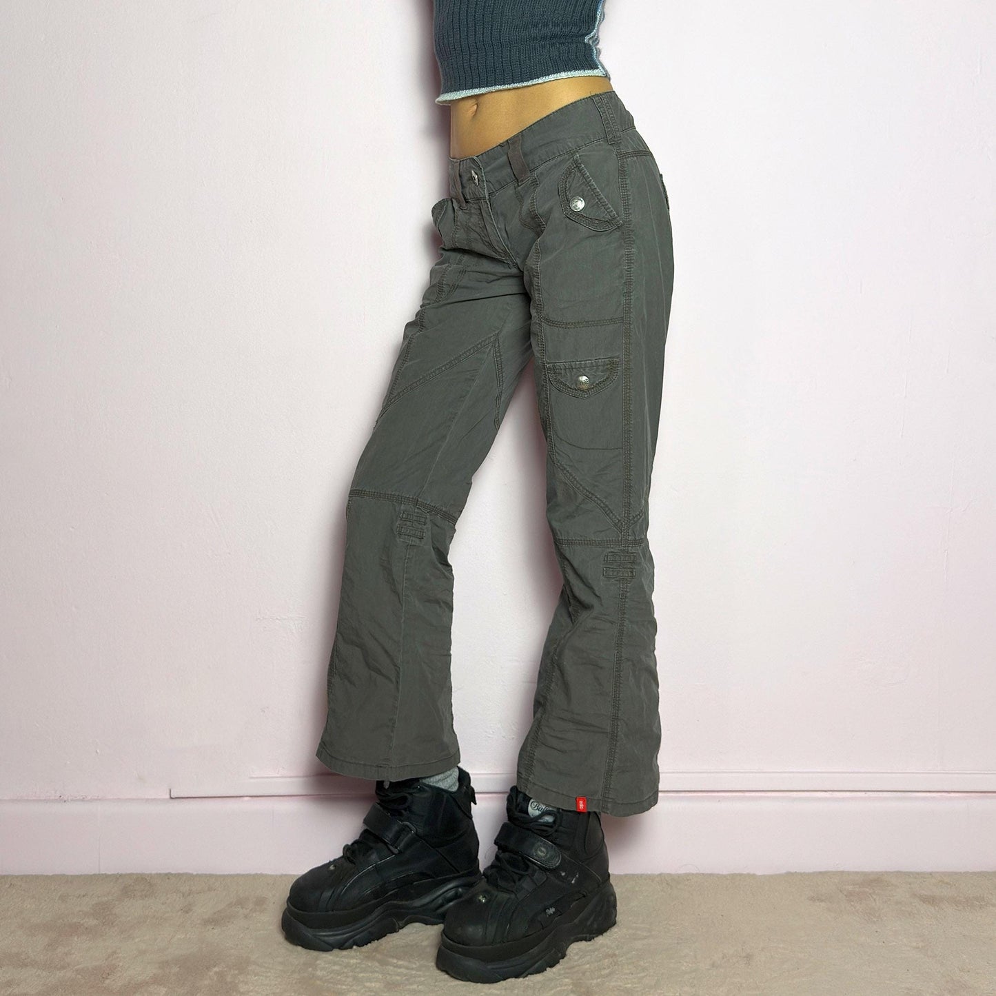 The Y2K Low-Rise Cargo Pants are a vintage 2000s piece featuring a washed gray tone with contrast stitching. Designed with a low-rise fit, button and zipper closure, and multiple cargo-style pockets, these pants have panel detailing and exposed seams that enhance their utilitarian aesthetic. The cropped straight-leg cut adds a modern touch, making them perfect for grunge and streetwear-inspired outfits. adult world shop
