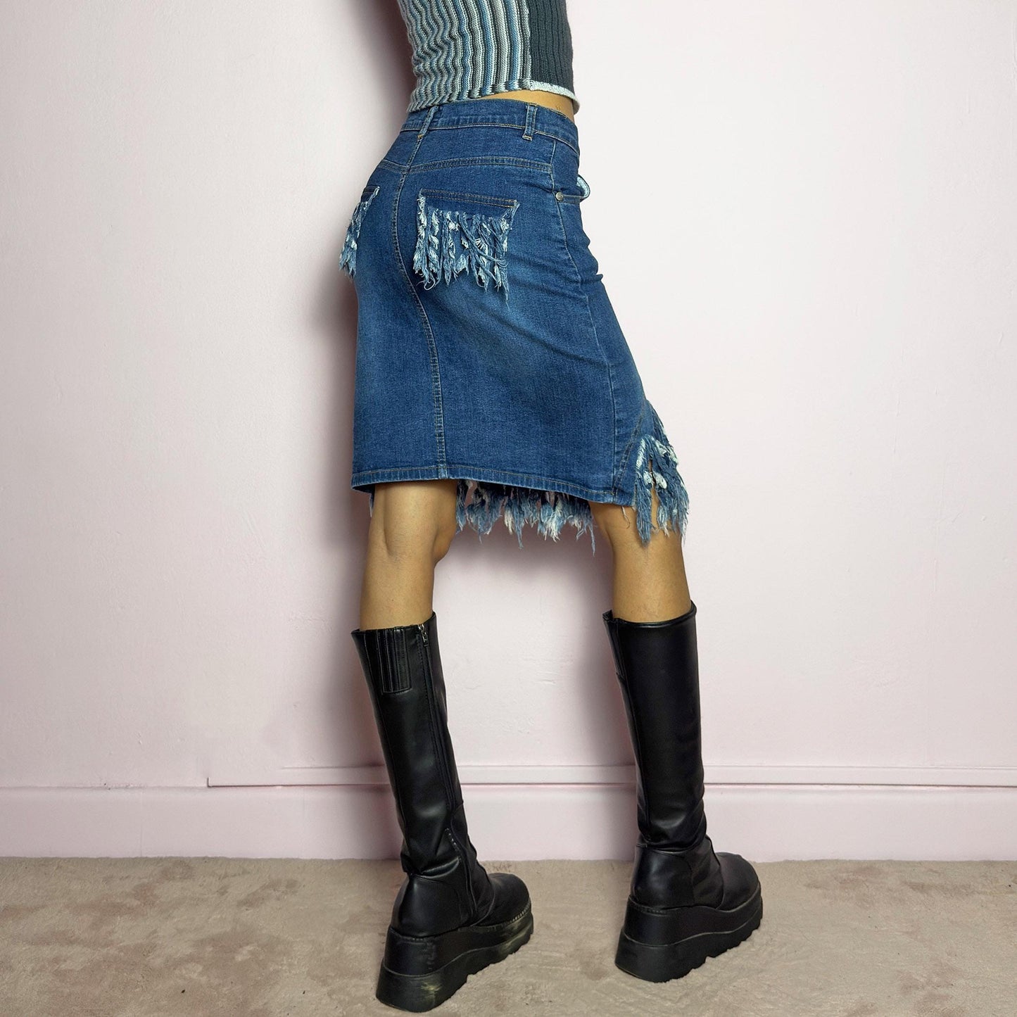 The Y2K Denim Distressed Midi Skirt is a vintage 2000s statement piece featuring a deconstructed look with dramatic distressed fringe details along the hem and back pockets. Designed with a stretchy structured denim fabric, a mid-rise fit, button and zipper closure, and pockets, this skirt blends edgy and casual aesthetics. adult world shop