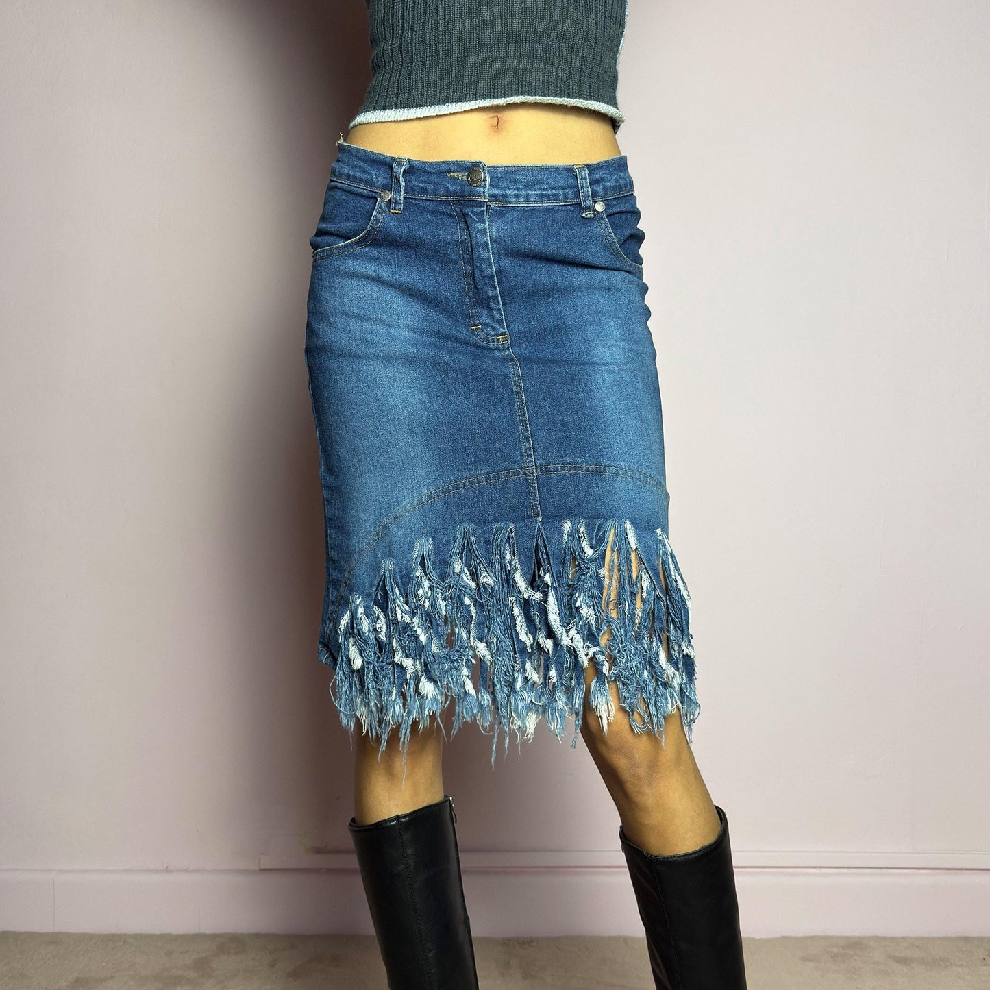 The Y2K Denim Distressed Midi Skirt is a vintage 2000s statement piece featuring a deconstructed look with dramatic distressed fringe details along the hem and back pockets. Designed with a stretchy structured denim fabric, a mid-rise fit, button and zipper closure, and pockets, this skirt blends edgy and casual aesthetics. adult world shop