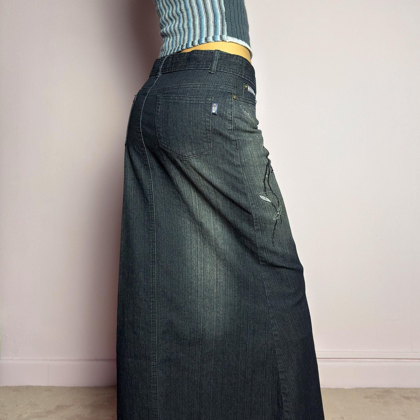 The Y2K Black Denim Maxi Skirt is a vintage 2000s piece featuring a sleek dark wash, rhinestone embellishments, and contrast stitching. Designed with a mid-rise fit and a stretchy denim fabric, it offers a structured silhouette that flares at the bottom. The skirt is finished with a button and zipper closure, pockets, and a removable belt for a customizable look. Perfect for casual streetwear outfits or an elegant night out. adult world shop
