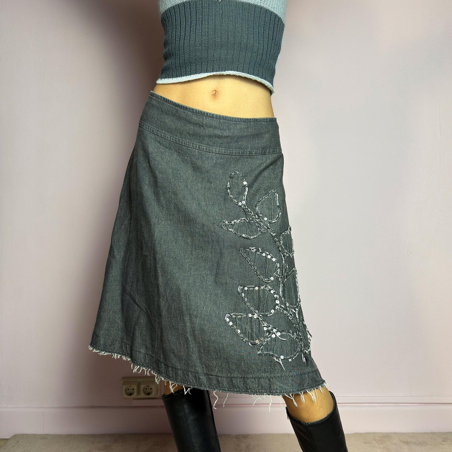 The Y2K Denim Gray Midi Skirt is a vintage 2000s piece featuring a faded gray wash, raw hem, and a unique floral sequin embroidery design. Crafted from structured denim, this skirt has an A-line silhouette that drapes effortlessly. Designed with a back zipper closure, the unfinished hem adds a deconstructed, edgy touch, making it perfect for casual outfits. adult world shop