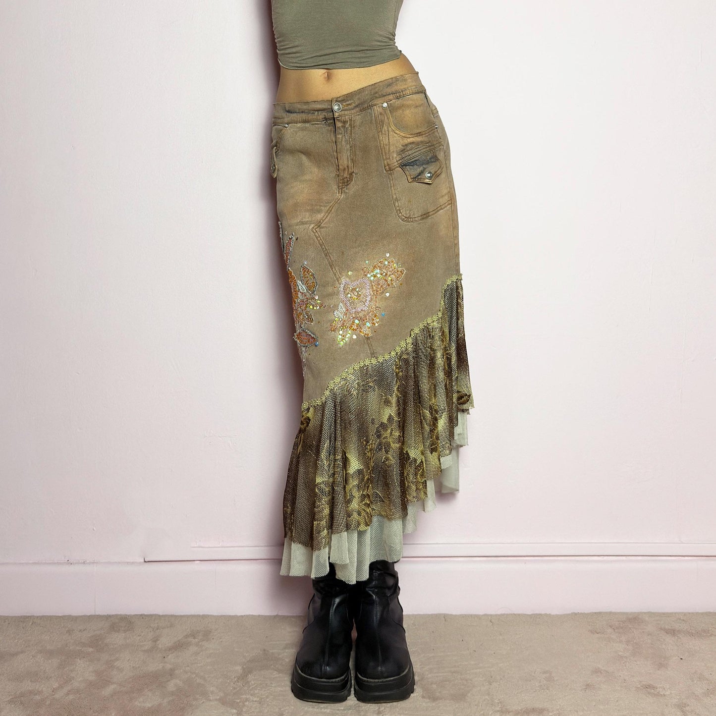 The Y2K Asymmetric Ruffle Midi Skirt is a vintage 2000s statement piece featuring a stretchy distressed tan denim base adorned with intricate beaded floral embroidery and sequin details. Designed with an asymmetric layered ruffle hem made of sheer lace and mesh with a metallic floral pattern, this skirt creates a dreamy, deconstructed aesthetic. It includes a button and zipper closure, belt loops, and pockets. adult world shop