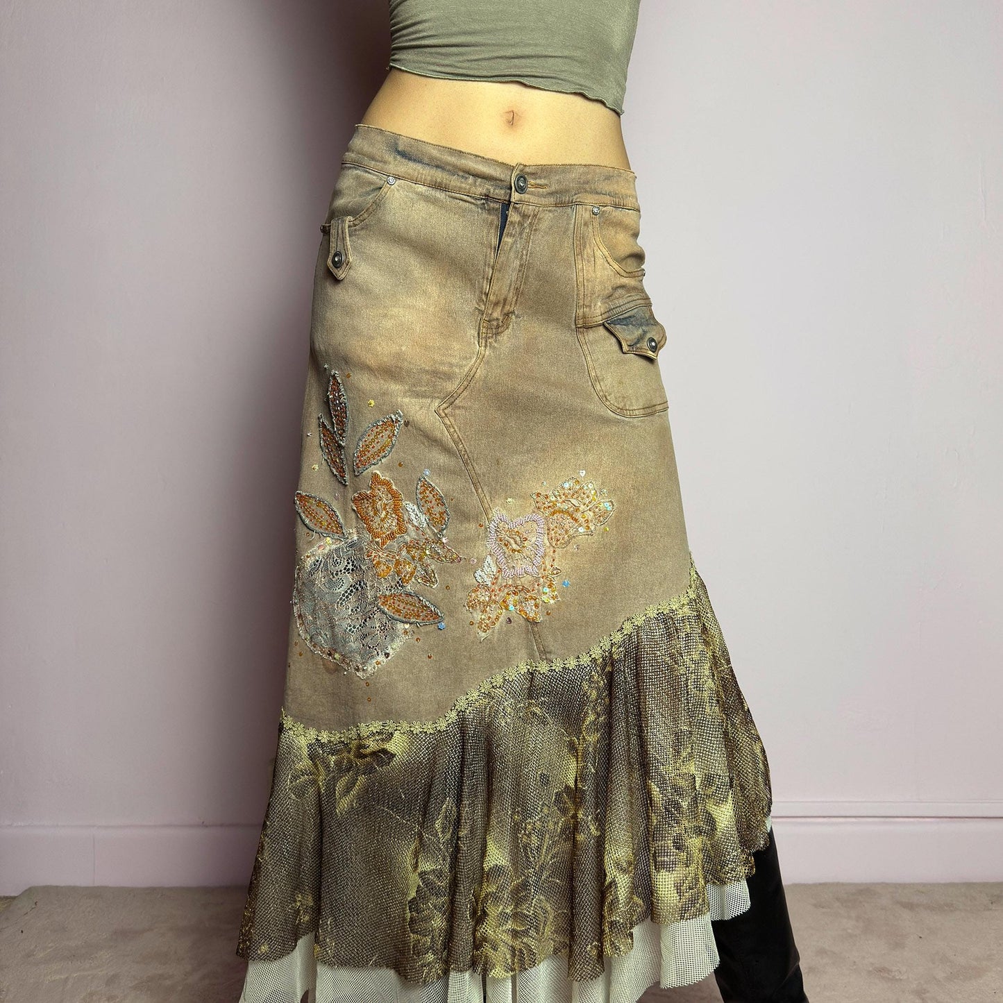 The Y2K Asymmetric Ruffle Midi Skirt is a vintage 2000s statement piece featuring a stretchy distressed tan denim base adorned with intricate beaded floral embroidery and sequin details. Designed with an asymmetric layered ruffle hem made of sheer lace and mesh with a metallic floral pattern, this skirt creates a dreamy, deconstructed aesthetic. It includes a button and zipper closure, belt loops, and pockets. adult world shop