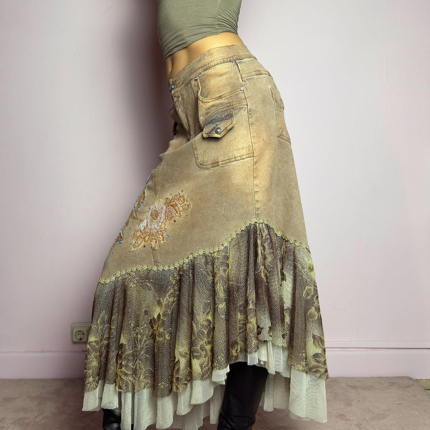 The Y2K Asymmetric Ruffle Midi Skirt is a vintage 2000s statement piece featuring a stretchy distressed tan denim base adorned with intricate beaded floral embroidery and sequin details. Designed with an asymmetric layered ruffle hem made of sheer lace and mesh with a metallic floral pattern, this skirt creates a dreamy, deconstructed aesthetic. It includes a button and zipper closure, belt loops, and pockets. adult world shop