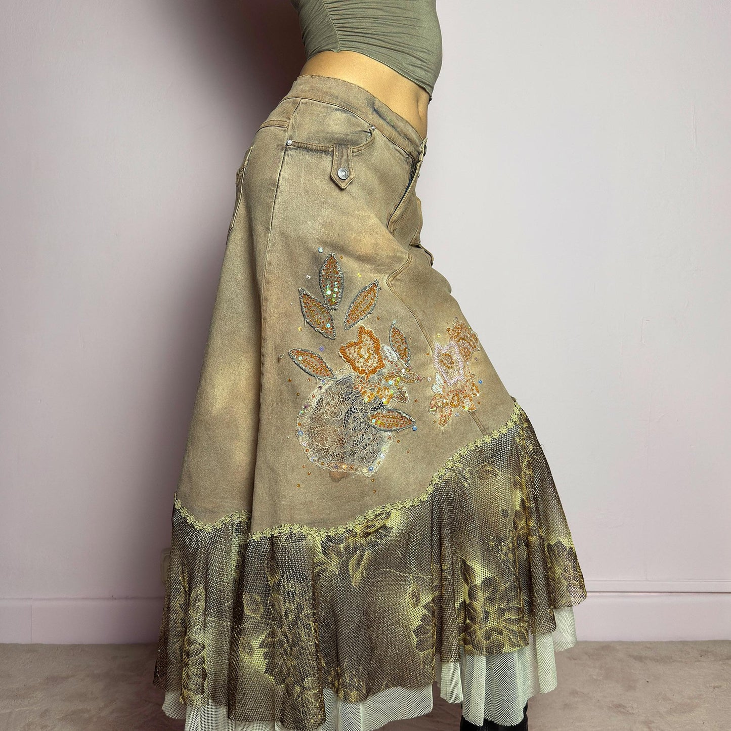 The Y2K Asymmetric Ruffle Midi Skirt is a vintage 2000s statement piece featuring a stretchy distressed tan denim base adorned with intricate beaded floral embroidery and sequin details. Designed with an asymmetric layered ruffle hem made of sheer lace and mesh with a metallic floral pattern, this skirt creates a dreamy, deconstructed aesthetic. It includes a button and zipper closure, belt loops, and pockets. adult world shop