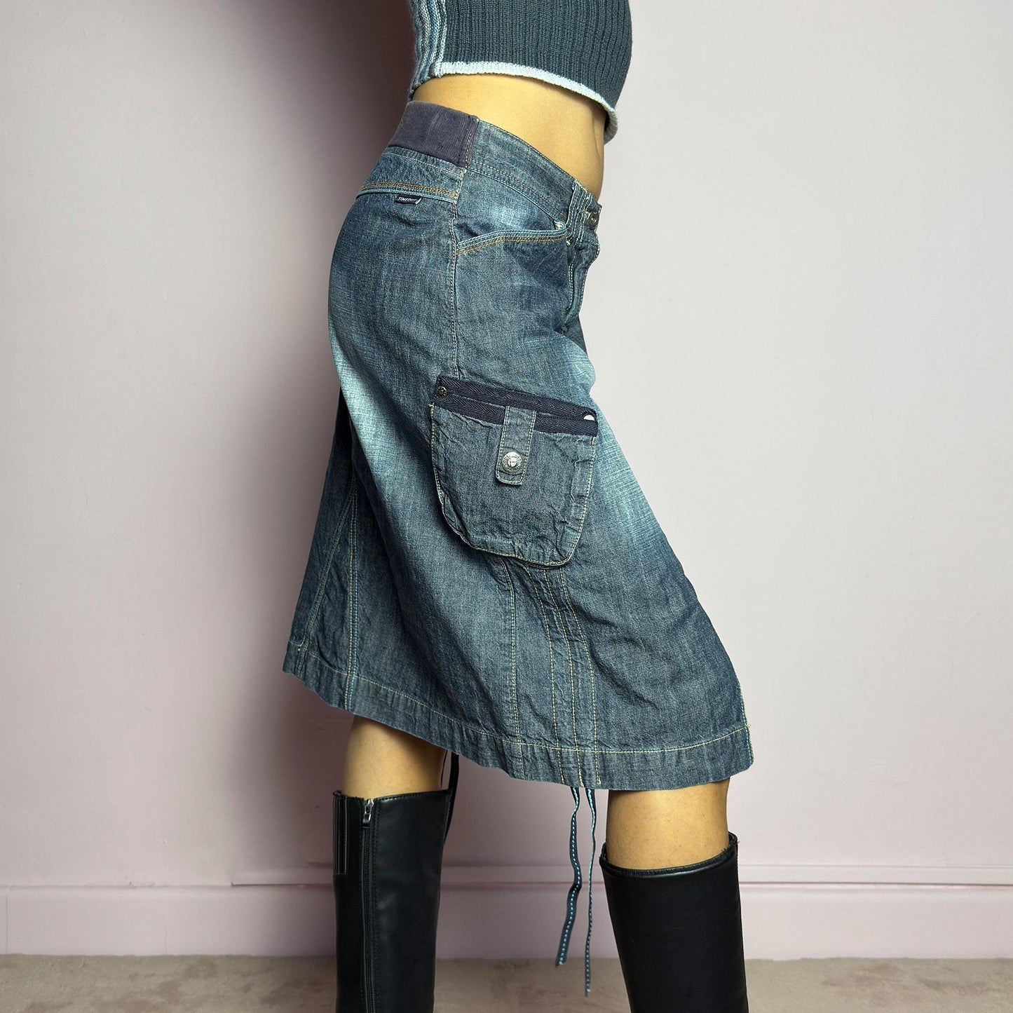 The Y2K Deconstructed Denim Midi Skirt is a vintage 2000s standout piece featuring a structured dark-wash denim fabric with contrast stitching. Designed with an asymmetrical front slit, utility-style cargo pockets, and adjustable drawstrings. Finished with a button and zipper closure, belt loops, and a mid-length silhouette, it's perfect for effortless grunge and utilitarian-inspired outfits. adult world shop