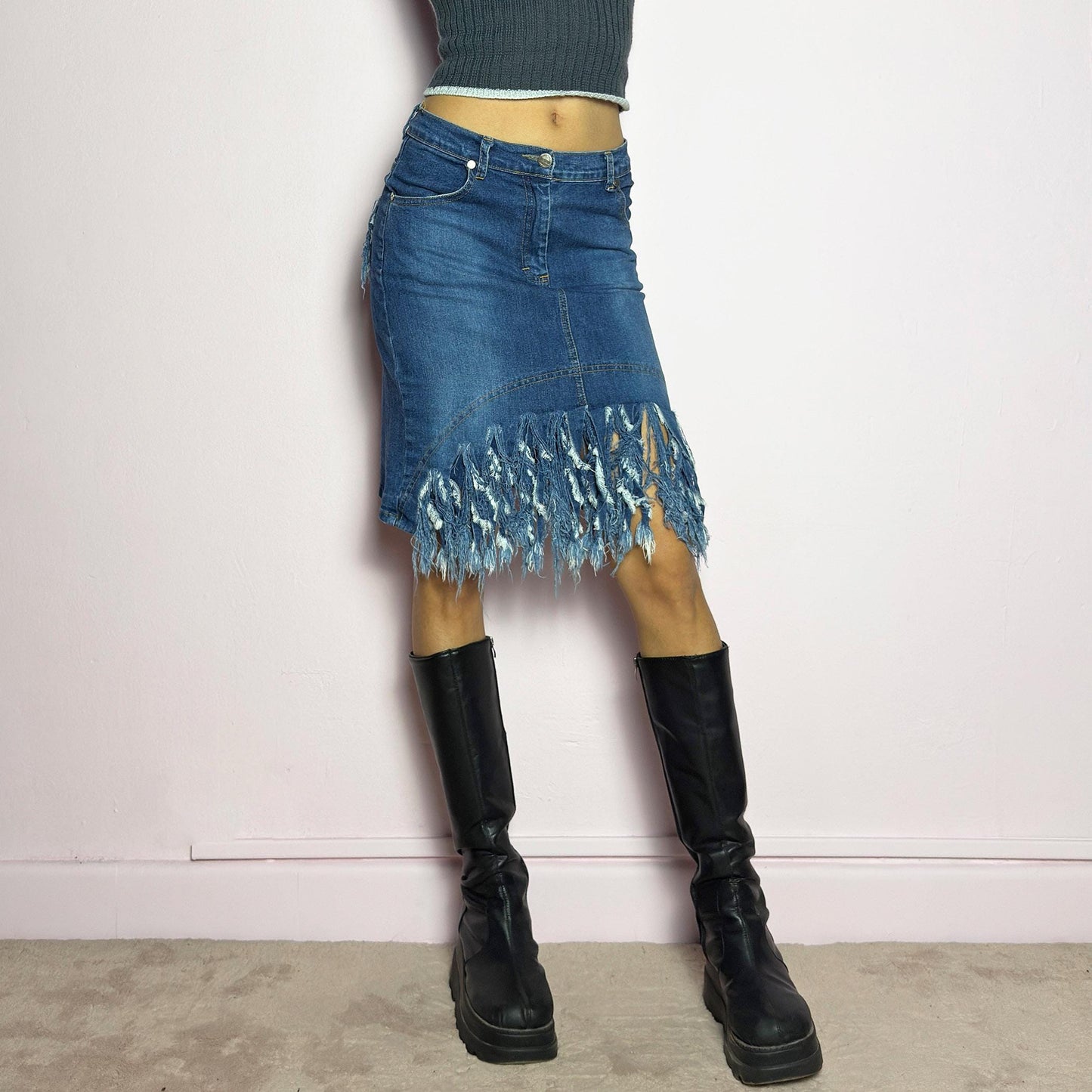 The Y2K Denim Distressed Midi Skirt is a vintage 2000s statement piece featuring a deconstructed look with dramatic distressed fringe details along the hem and back pockets. Designed with a stretchy structured denim fabric, a mid-rise fit, button and zipper closure, and pockets, this skirt blends edgy and casual aesthetics. adult world shop