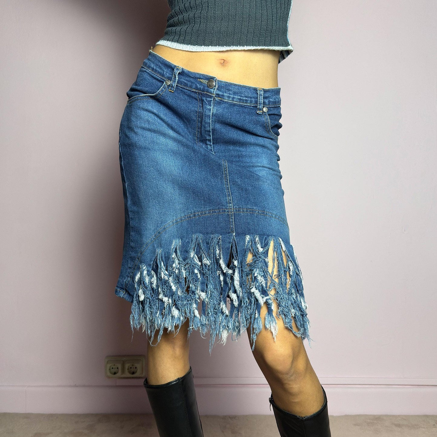 The Y2K Denim Distressed Midi Skirt is a vintage 2000s statement piece featuring a deconstructed look with dramatic distressed fringe details along the hem and back pockets. Designed with a stretchy structured denim fabric, a mid-rise fit, button and zipper closure, and pockets, this skirt blends edgy and casual aesthetics. adult world shop
