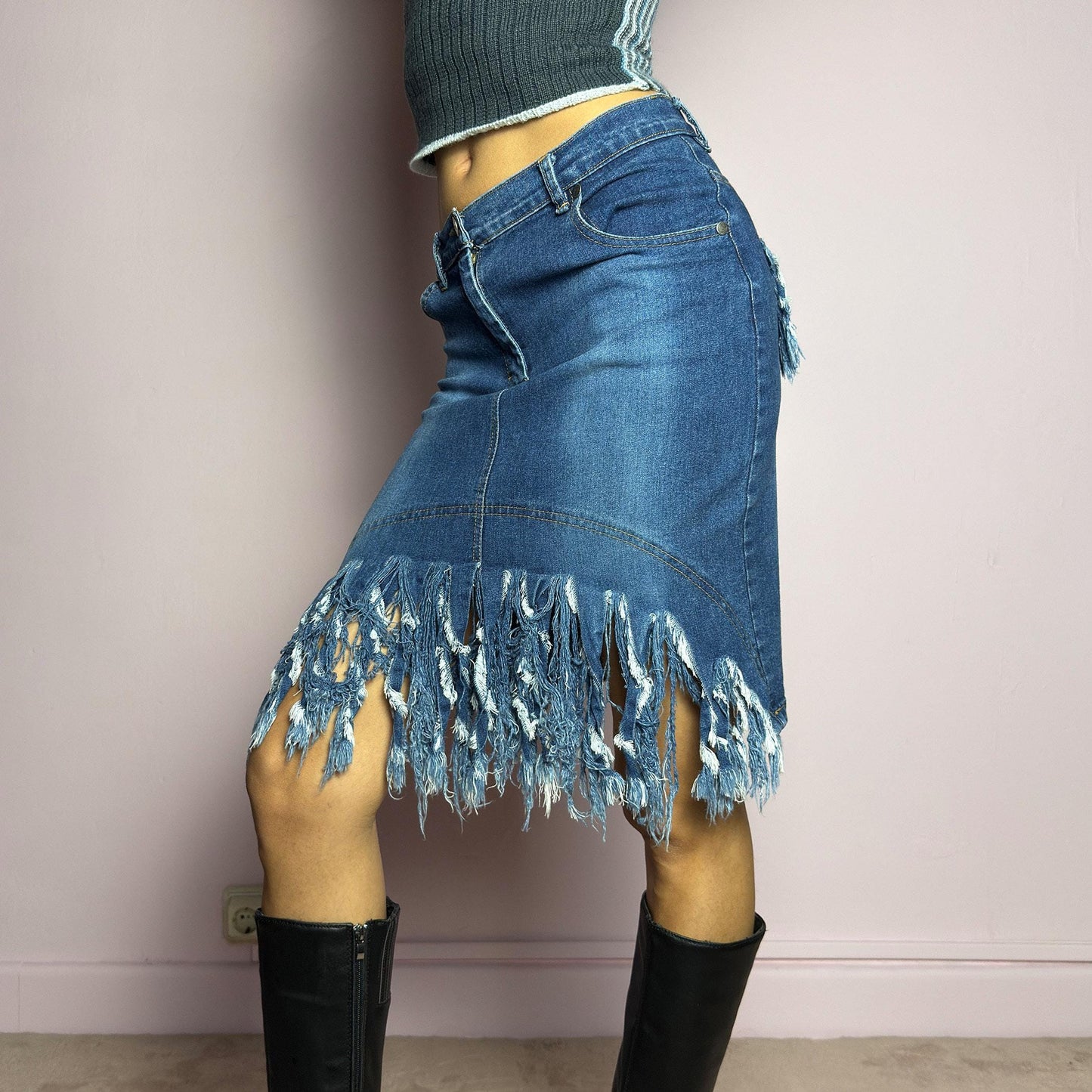 The Y2K Denim Distressed Midi Skirt is a vintage 2000s statement piece featuring a deconstructed look with dramatic distressed fringe details along the hem and back pockets. Designed with a stretchy structured denim fabric, a mid-rise fit, button and zipper closure, and pockets, this skirt blends edgy and casual aesthetics. adult world shop