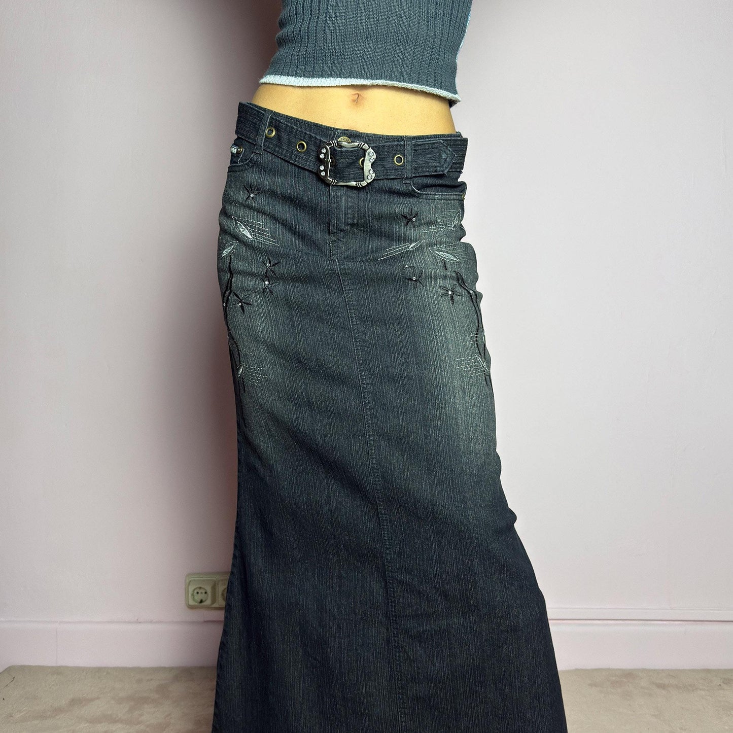 The Y2K Black Denim Maxi Skirt is a vintage 2000s piece featuring a sleek dark wash, rhinestone embellishments, and contrast stitching. Designed with a mid-rise fit and a stretchy denim fabric, it offers a structured silhouette that flares at the bottom. The skirt is finished with a button and zipper closure, pockets, and a removable belt for a customizable look. Perfect for casual streetwear outfits or an elegant night out. adult world shop