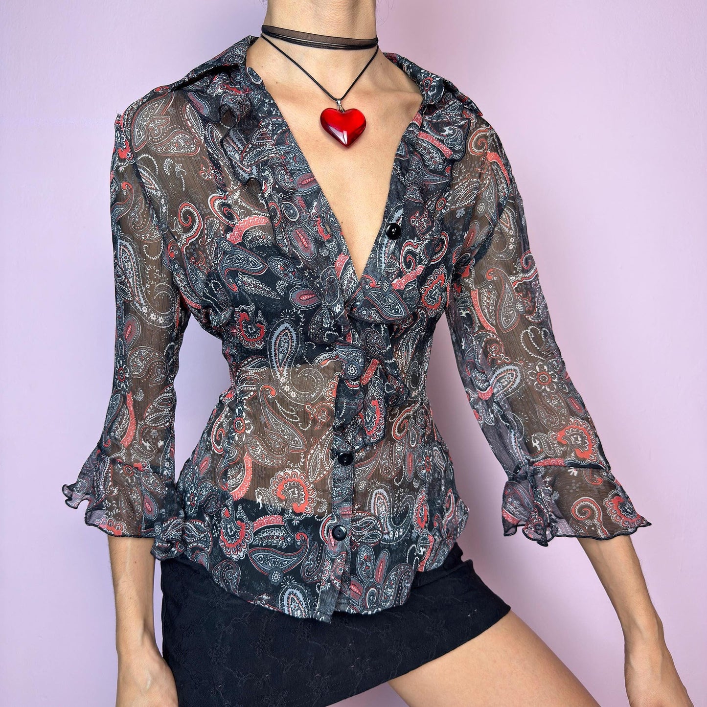 The Vintage 90s Black Ruffle Sheer Blouse features a semi-sheer fabric with an allover paisley print in red, grey, and white tones. This fitted silhouette includes three-quarter bell sleeves with flared ruffle cuffs, a romantic ruffled collar, and a front button-up closure. The lightweight fabric makes it perfect for summer layering or wearing on its own for a sleek, sexy vibe. Great vintage condition. adult world shop