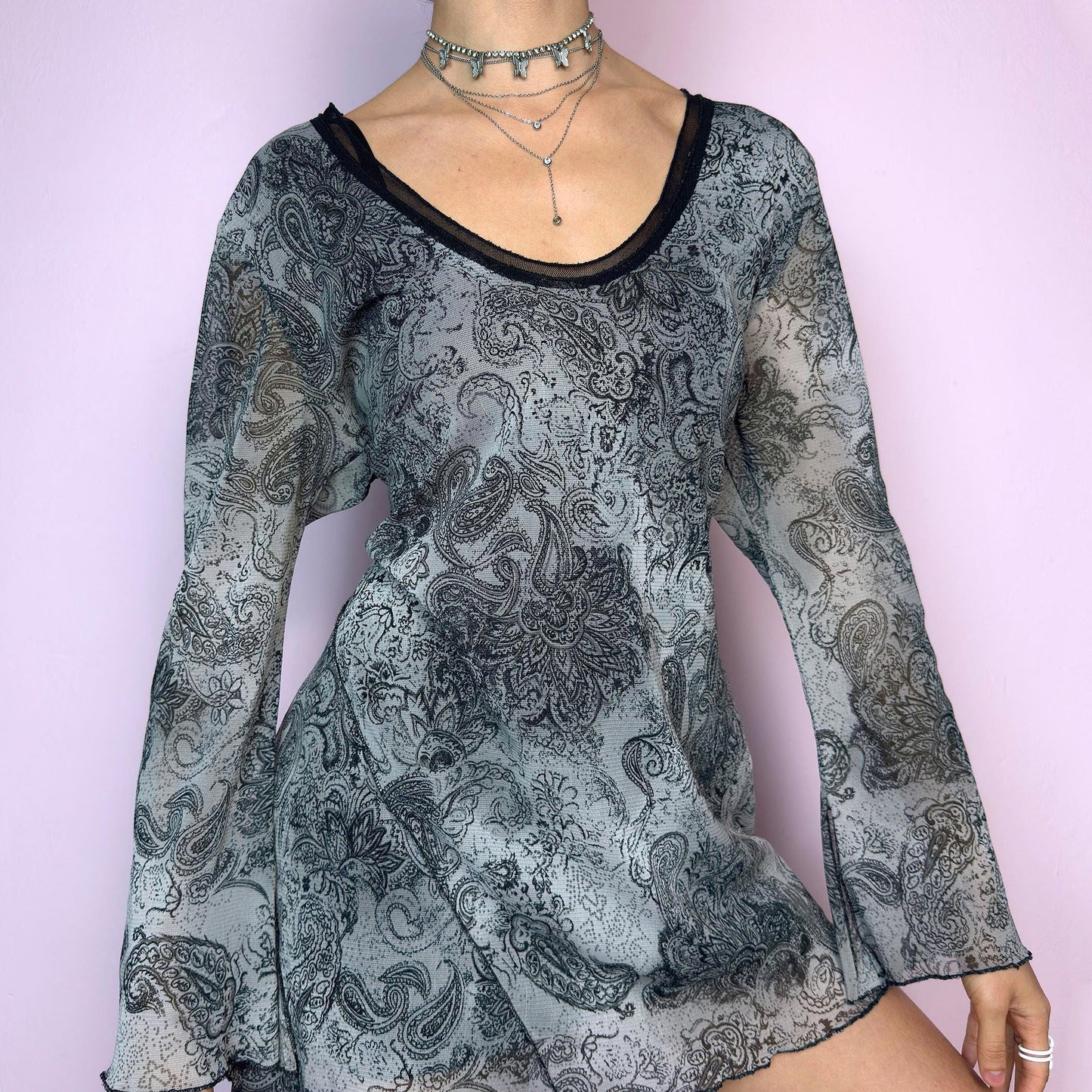 The Y2K Gray Asymmetric Mesh Top is a vintage 2000s long sleeve blouse featuring an all-over black and grey paisley print. Crafted from a lightweight mesh fabric, it has a whimsical, fairy-inspired silhouette with subtle bell sleeves and an asymmetric hemline that adds dreamy movement. Perfect for casual spring or festival looks. Excellent vintage condition. adult world shop