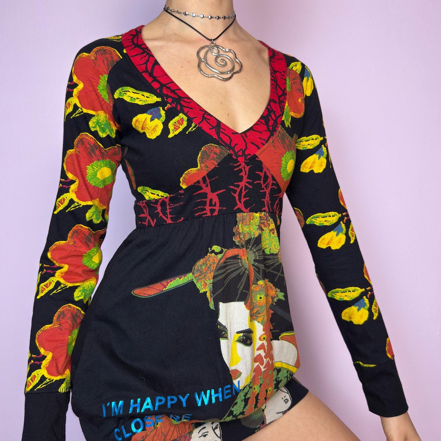 The Y2K Desigual Graphic Print Top is a rare vintage 2000s long sleeve top by Desigual featuring an eclectic mix of prints and bold colors. This artsy piece combines floral motifs, illustrated portraits, and poetic text layered over a stretchy black base. Designed with a deep V-neckline and empire waist for a flattering silhouette. The bottom hem is subtly gathered for a closer fit, giving it a mini dress vibe. Includes hidden side pockets. adult world shop