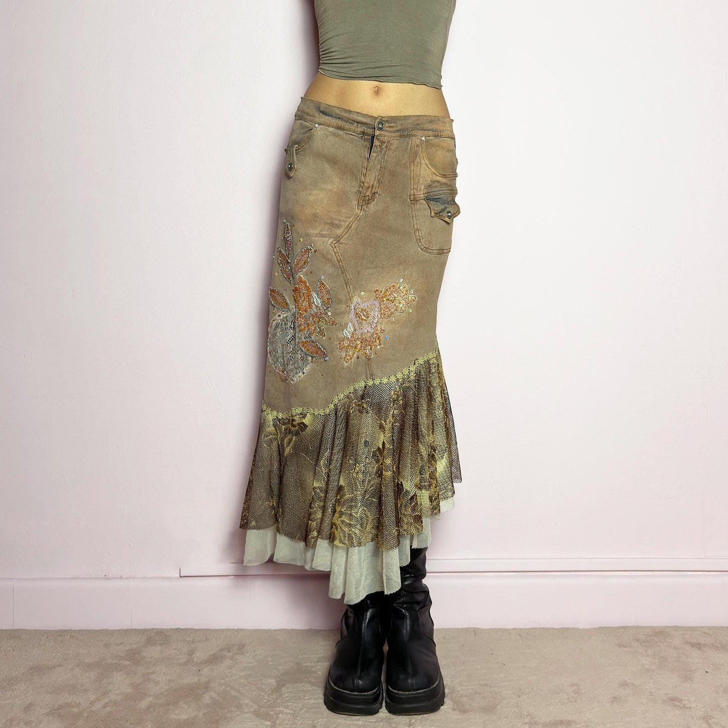The Y2K Asymmetric Ruffle Midi Skirt is a vintage 2000s statement piece featuring a stretchy distressed tan denim base adorned with intricate beaded floral embroidery and sequin details. Designed with an asymmetric layered ruffle hem made of sheer lace and mesh with a metallic floral pattern, this skirt creates a dreamy, deconstructed aesthetic. It includes a button and zipper closure, belt loops, and pockets. adult world shop