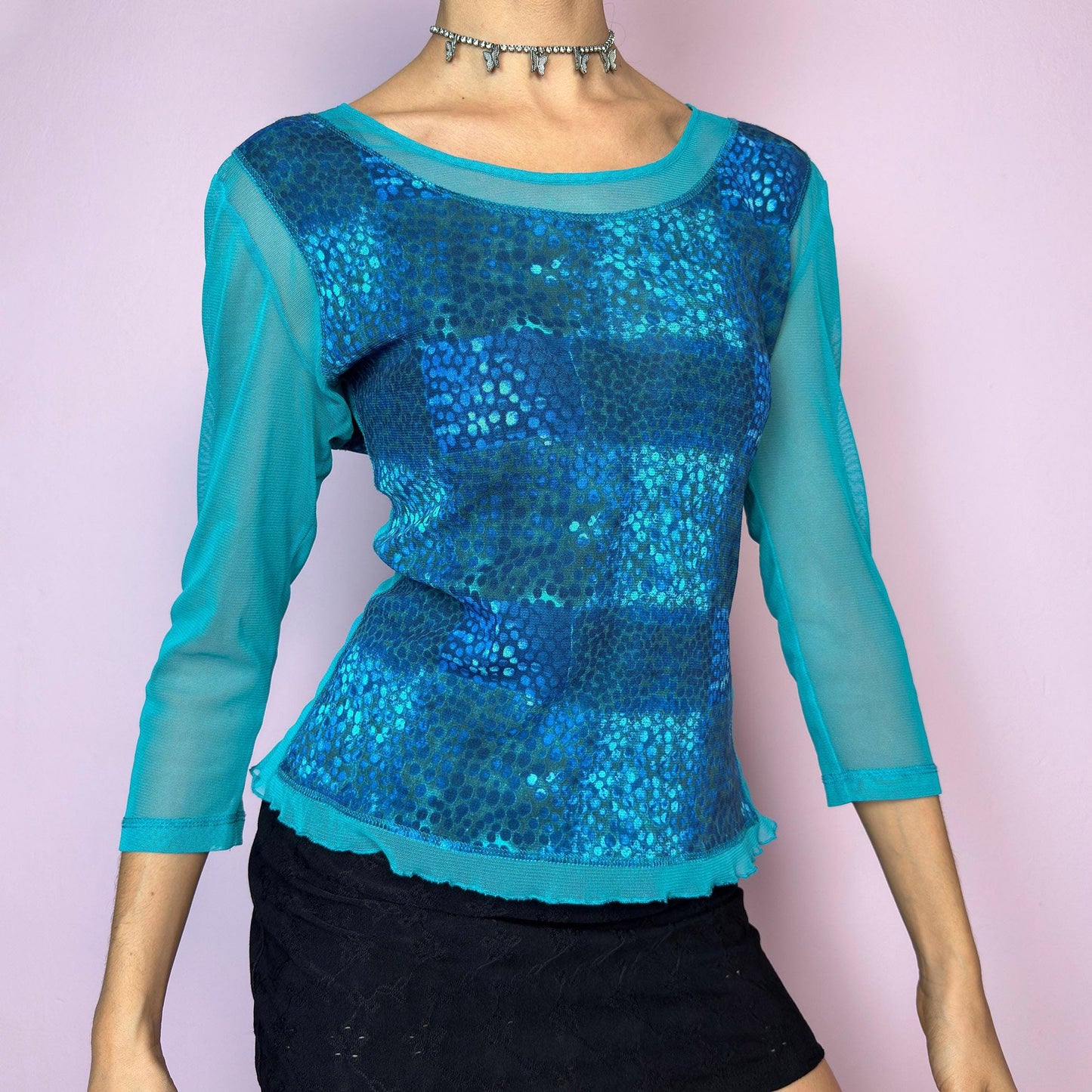 The Y2K Blue Patchwork Mesh Top is a vintage 2000s blue mesh top featuring a striking patchwork-style abstract print in shades of turquoise and electric blue. Designed with semi-sheer mesh sleeves and body, it has a fitted silhouette and three-quarter length sleeves. Lightweight and stretchy, this top is perfect for layering in rave, festival, or cyber looks. Excellent vintage condition.