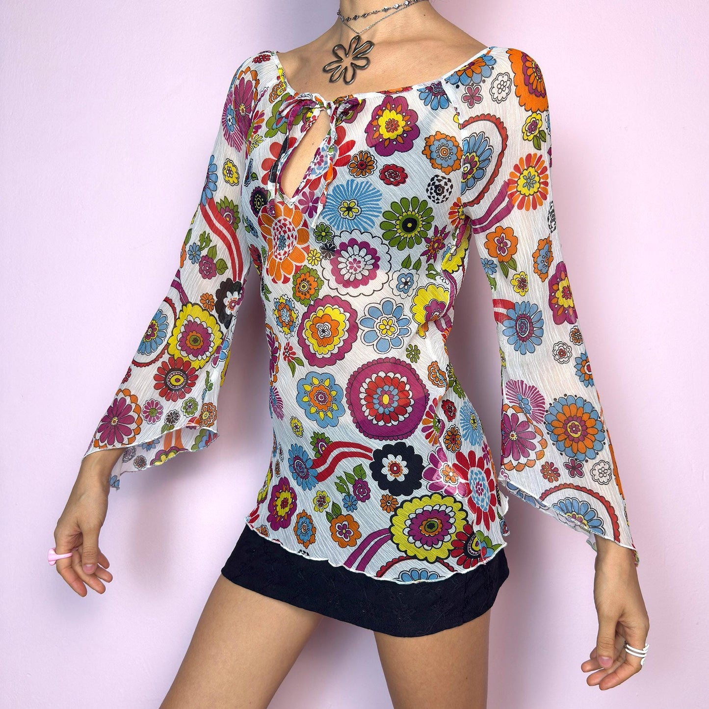 The Y2K White Floral Tie Front Top is a vintage 2000s piece bursting with color and retro charm. Features a vibrant multicolor 60s-inspired flower print on lightweight, slightly sheer pleated fabric. Designed with a flattering scoop neckline, keyhole front with tie closure, and flared bell sleeves. Stretchy and breathable – perfect for summer, festivals, or hippie styles. Great vintage condition.