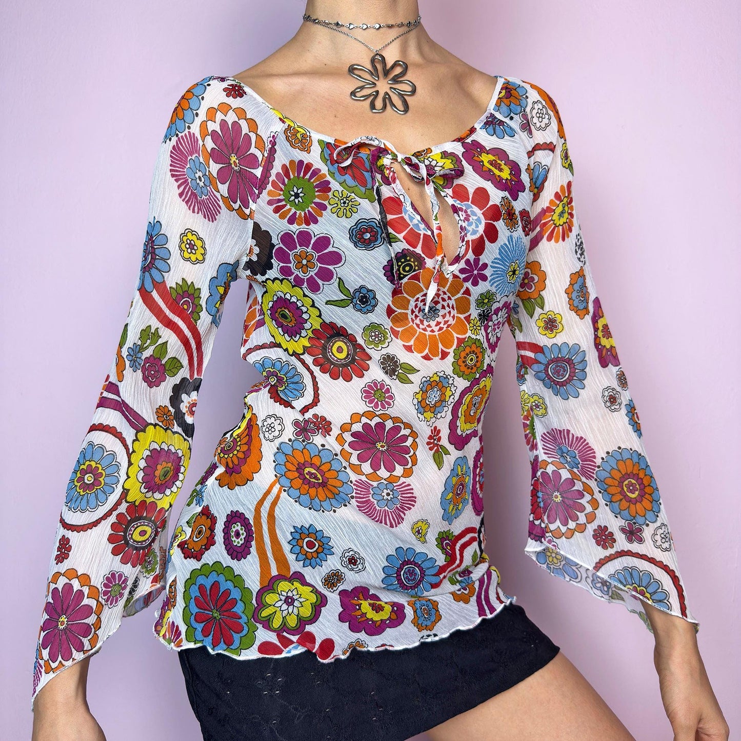 The Y2K White Floral Tie Front Top is a vintage 2000s piece bursting with color and retro charm. Features a vibrant multicolor 60s-inspired flower print on lightweight, slightly sheer pleated fabric. Designed with a flattering scoop neckline, keyhole front with tie closure, and flared bell sleeves. Stretchy and breathable – perfect for summer, festivals, or hippie styles. Great vintage condition.