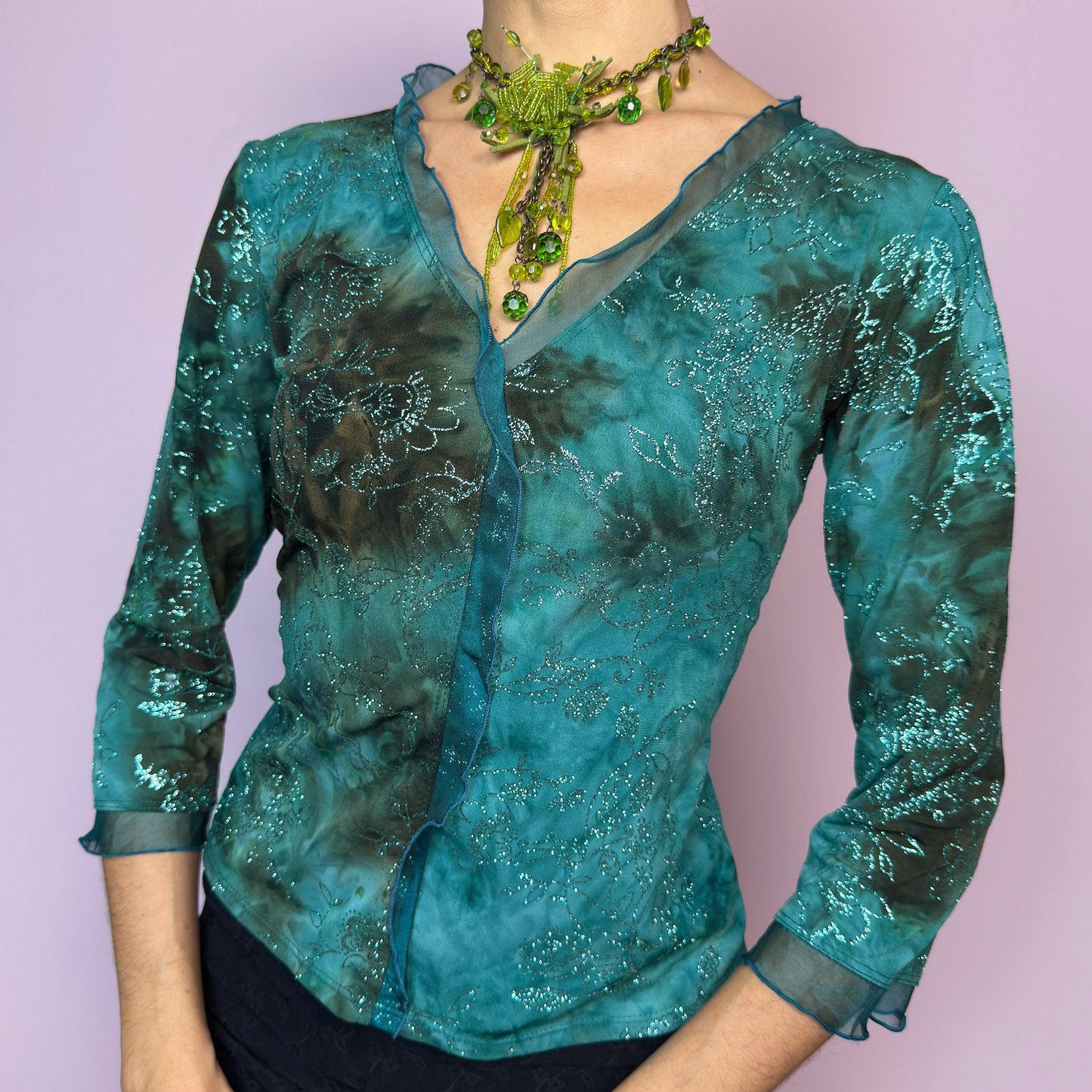 The Vintage 90s Teal Tie Dye Glitter Top is a stunning piece featuring a soft ruffle trim along the V-neckline, a dreamy tie-dye wash in teal and mossy dark tones, and shimmering metallic floral glitter details. Designed with three-quarter sleeves and delicate chiffon-like hems. The fabric is stretchy for a comfortable, flattering fit. This romantic blouse captures the essence of fairy grunge, boho nights, and witchy glam styling. Perfect for adding a touch of ethereal magic to your wardrobe.