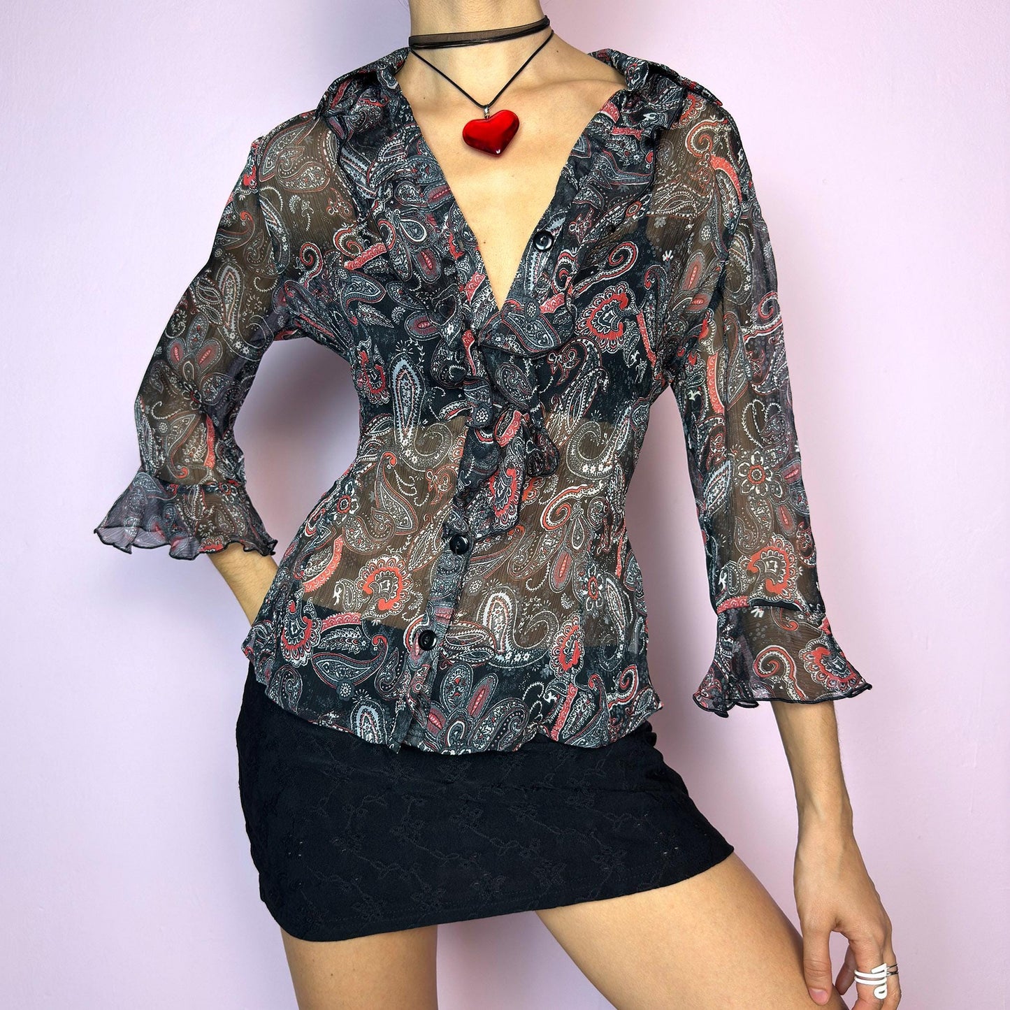 The Vintage 90s Black Ruffle Sheer Blouse features a semi-sheer fabric with an allover paisley print in red, grey, and white tones. This fitted silhouette includes three-quarter bell sleeves with flared ruffle cuffs, a romantic ruffled collar, and a front button-up closure. The lightweight fabric makes it perfect for summer layering or wearing on its own for a sleek, sexy vibe. Great vintage condition. adult world shop