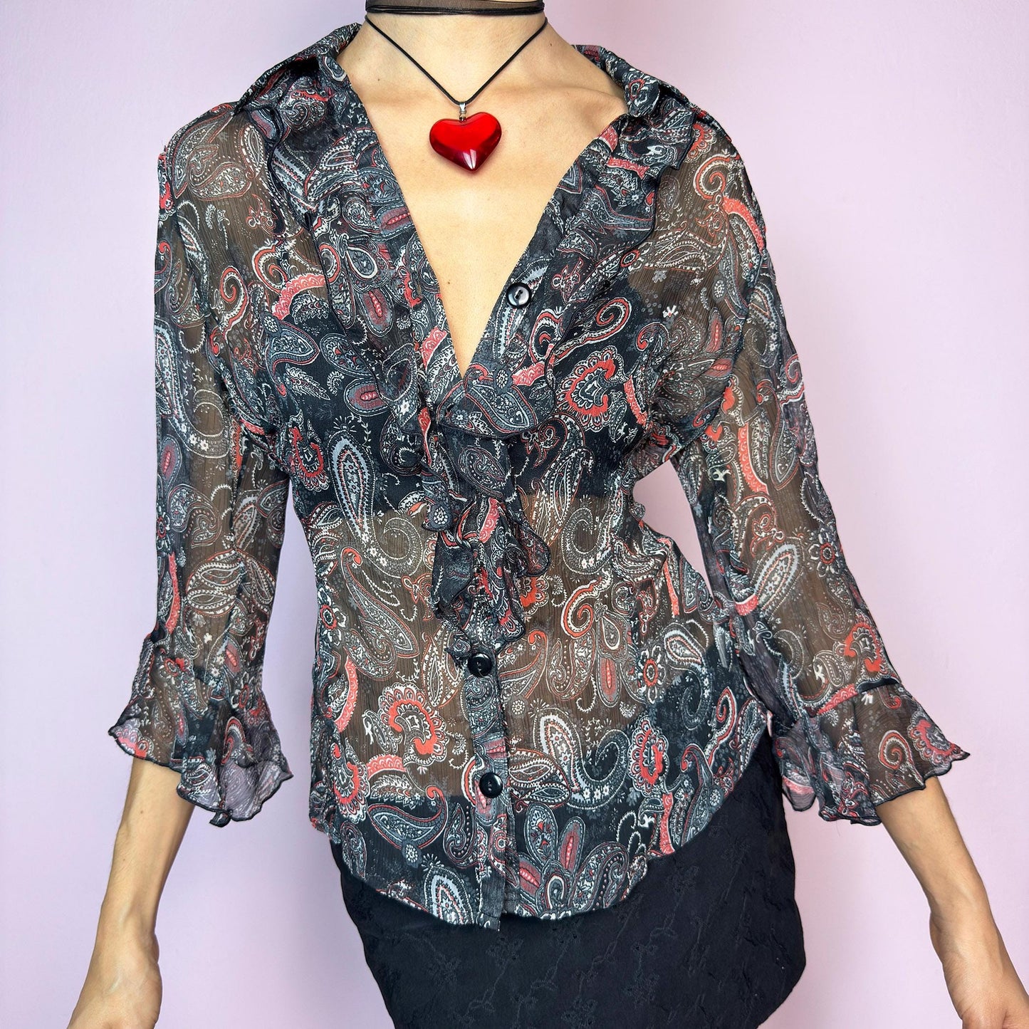 The Vintage 90s Black Ruffle Sheer Blouse features a semi-sheer fabric with an allover paisley print in red, grey, and white tones. This fitted silhouette includes three-quarter bell sleeves with flared ruffle cuffs, a romantic ruffled collar, and a front button-up closure. The lightweight fabric makes it perfect for summer layering or wearing on its own for a sleek, sexy vibe. Great vintage condition. adult world shop