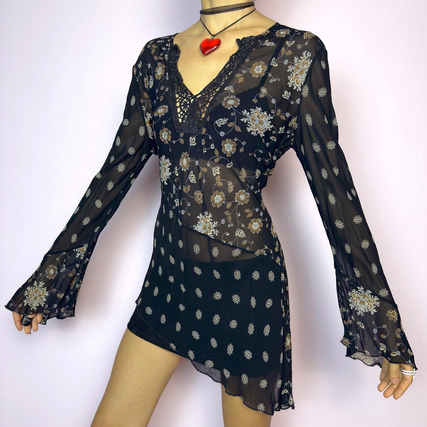 The Y2K Asymmetric Bell Sleeve Top is a vintage 2000s piece featuring a sheer black floral print with an asymmetric hem and dramatic bell sleeves for a flowy silhouette. The deep V-neckline is accented with crochet lace trim for a romantic touch. Lightweight and breathable, it’s perfect for summer layering or festival styling. Includes long sleeves with a bell flare for an elegant witchy vibe. Great vintage condition. adult world shop
