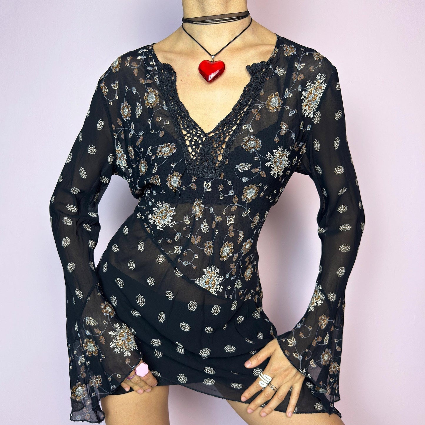 The Y2K Asymmetric Bell Sleeve Top is a vintage 2000s piece featuring a sheer black floral print with an asymmetric hem and dramatic bell sleeves for a flowy silhouette. The deep V-neckline is accented with crochet lace trim for a romantic touch. Lightweight and breathable, it’s perfect for summer layering or festival styling. Includes long sleeves with a bell flare for an elegant witchy vibe. Great vintage condition. adult world shop