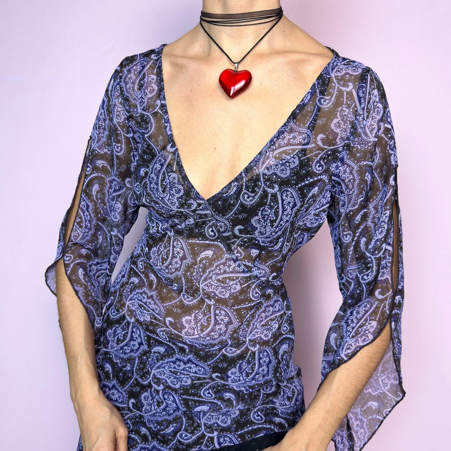 The Y2K Black Sheer Paisley Blouse is a vintage 2000s sheer paisley print blouse in black and purple tones. Features an asymmetrical hemline, open slit bell sleeves, and a flattering deep V-neckline with a tie-back detail to cinch the waist. Made from lightweight fabric for a breezy fit. Perfect for summer layering, night-out looks, or festival styling. Great vintage condition. adult world shop