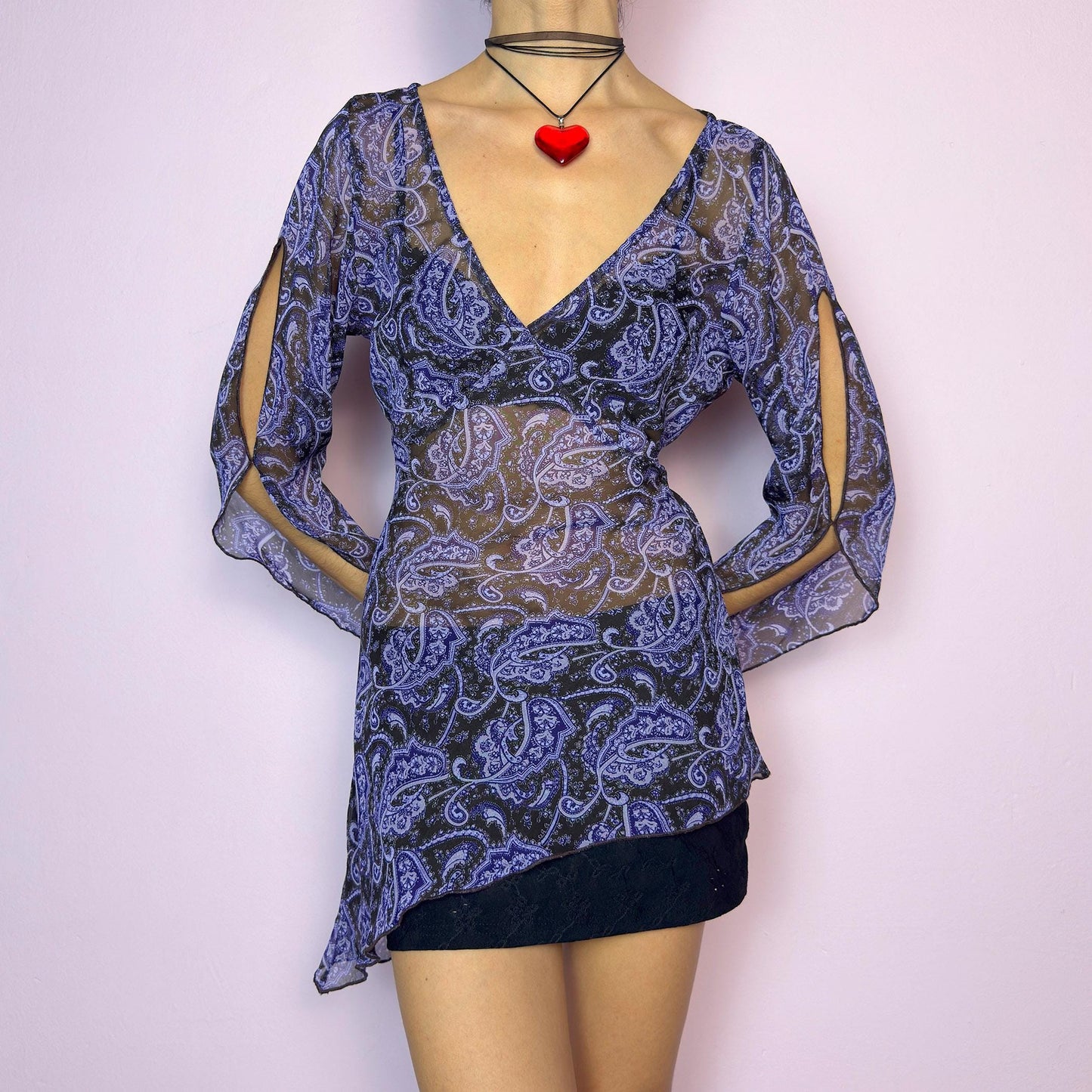 The Y2K Black Sheer Paisley Blouse is a vintage 2000s sheer paisley print blouse in black and purple tones. Features an asymmetrical hemline, open slit bell sleeves, and a flattering deep V-neckline with a tie-back detail to cinch the waist. Made from lightweight fabric for a breezy fit. Perfect for summer layering, night-out looks, or festival styling. Great vintage condition. adult world shop