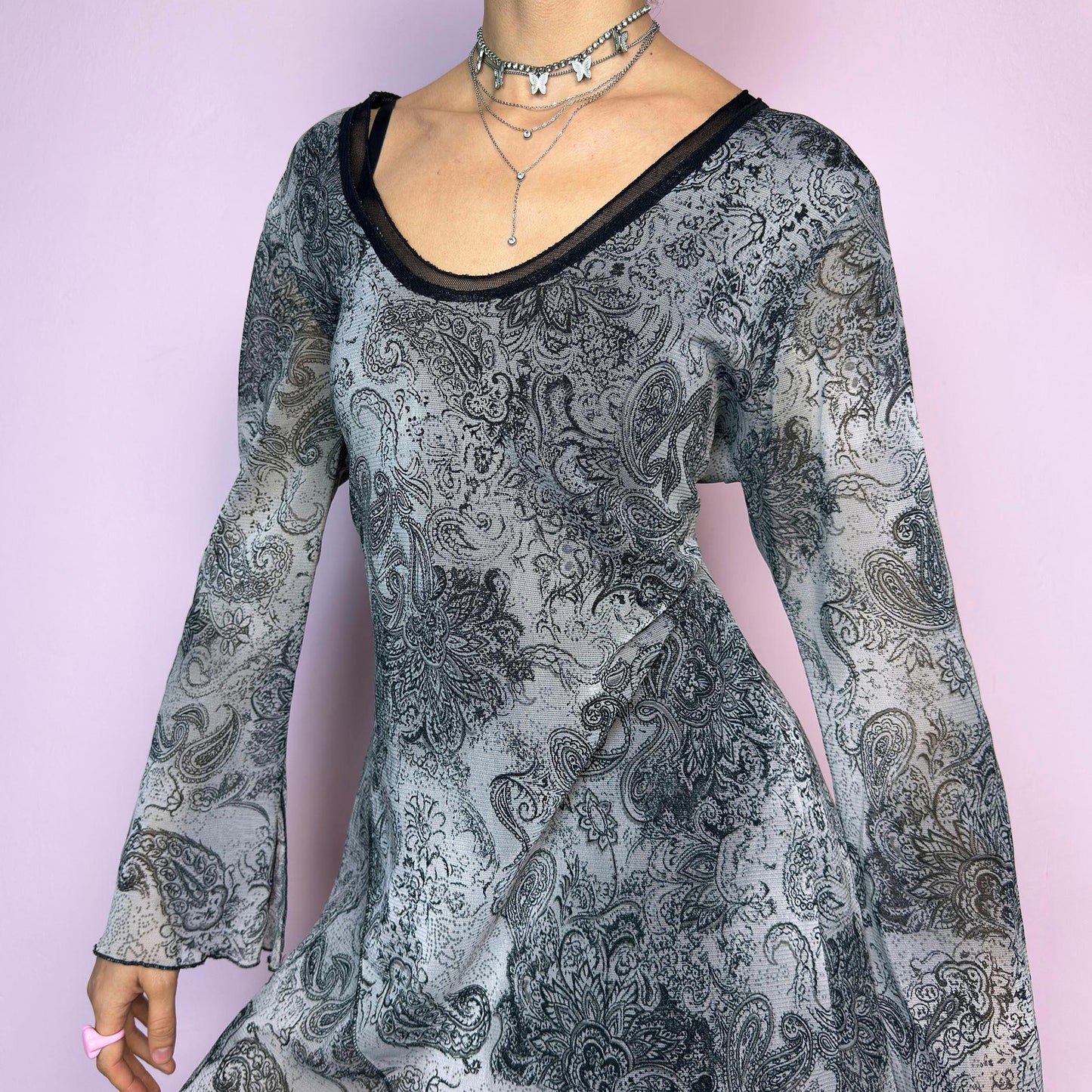 The Y2K Gray Asymmetric Mesh Top is a vintage 2000s long sleeve blouse featuring an all-over black and grey paisley print. Crafted from a lightweight mesh fabric, it has a whimsical, fairy-inspired silhouette with subtle bell sleeves and an asymmetric hemline that adds dreamy movement. Perfect for casual spring or festival looks. Excellent vintage condition. adult world shop