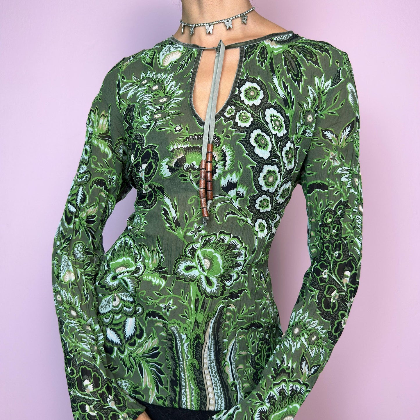 The Y2K Green Boho Tie Front Top is a vintage 2000s piece featuring an abstract floral print on lightweight, semi-sheer fabric. Designed with a front keyhole neckline and adjustable tie adorned with wooden bead details. The long sleeves and breathable material make it comfy and flattering. A unique top perfect for summer layering, festival outfits, fairy grunge, or earthy boho styles.