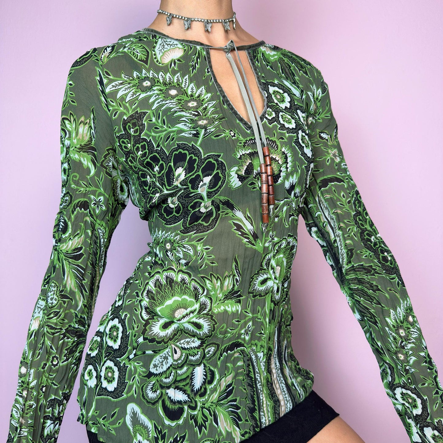 The Y2K Green Boho Tie Front Top is a vintage 2000s piece featuring an abstract floral print on lightweight, semi-sheer fabric. Designed with a front keyhole neckline and adjustable tie adorned with wooden bead details. The long sleeves and breathable material make it comfy and flattering. A unique top perfect for summer layering, festival outfits, fairy grunge, or earthy boho styles.