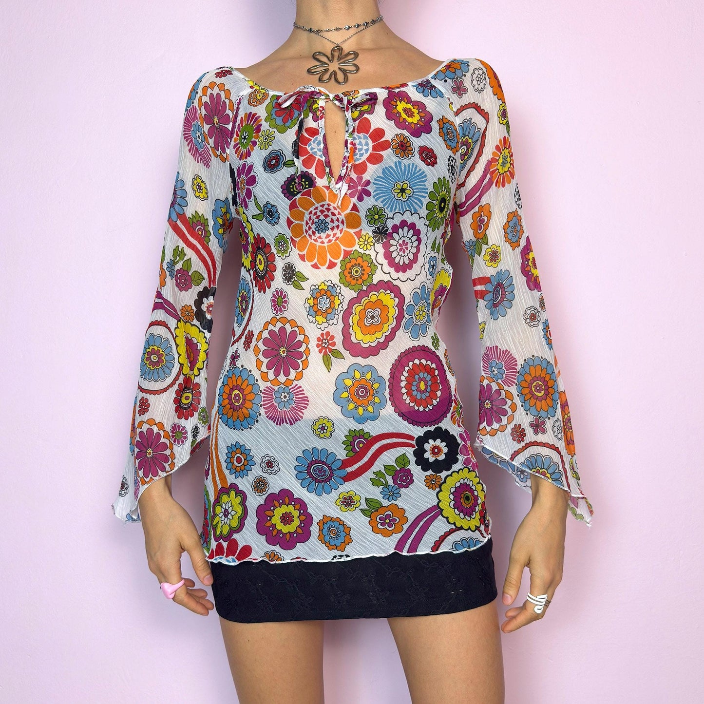 The Y2K White Floral Tie Front Top is a vintage 2000s piece bursting with color and retro charm. Features a vibrant multicolor 60s-inspired flower print on lightweight, slightly sheer pleated fabric. Designed with a flattering scoop neckline, keyhole front with tie closure, and flared bell sleeves. Stretchy and breathable – perfect for summer, festivals, or hippie styles. Great vintage condition.