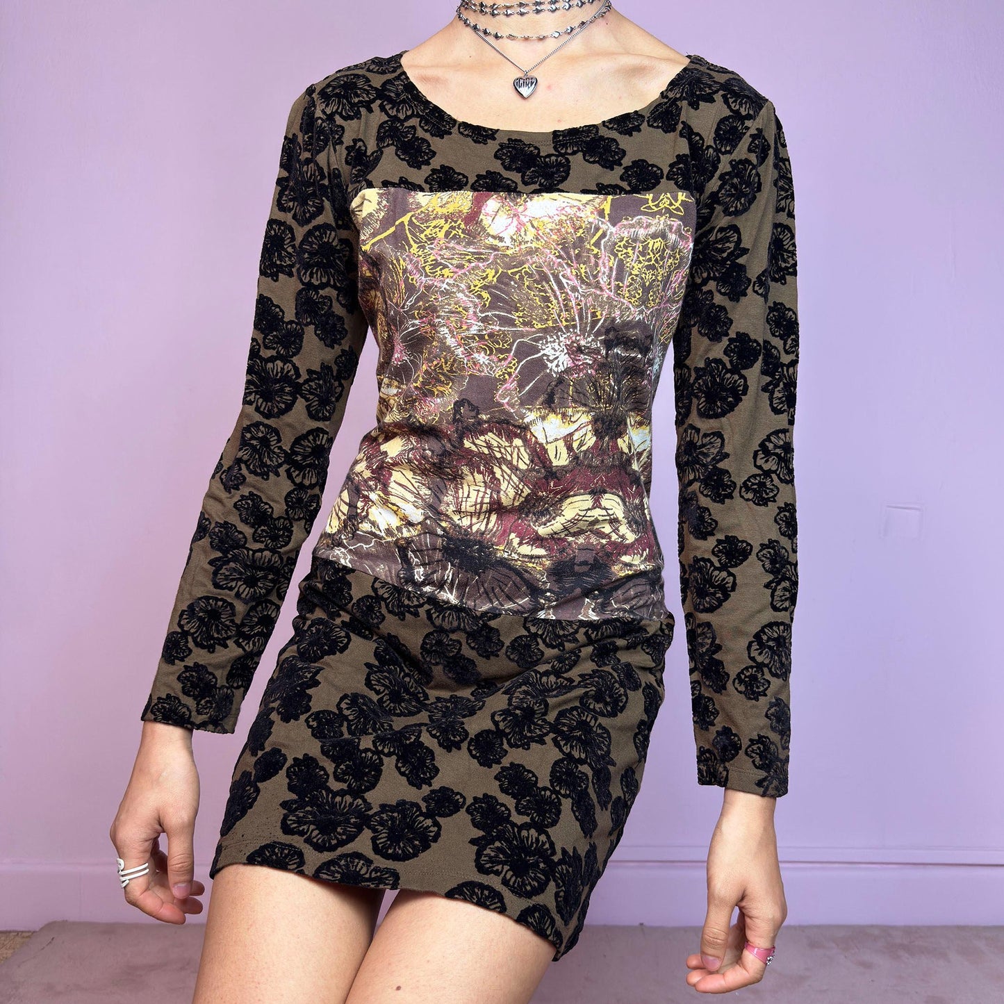 The Y2K Printed Velvet Mini Dress is a striking and artsy vintage piece from the 2000s. This stretchy mini dress features a unique mixed-media design with an abstract print panel at the chest, contrasted by a brown base covered in bold black velvet flocked florals. With its long sleeves, slightly flared hem, and figure-hugging silhouette, it's perfect for maximalist layering or making a statement on its own. A rare cyber-romantic gem for experimental, alt, or fairy-grunge wardrobes.