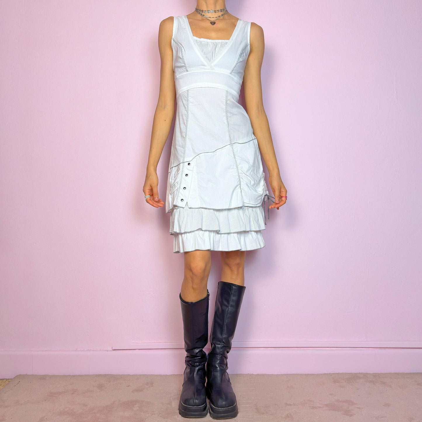This vintage 2000s white sleeveless dress features a square neckline and sculpted panel seams. Made from a stretchy fabric, it includes a side zipper and falls to a midi length. Grommet hardware and a ruched drawstring side add a utilitarian twist, while the double-tiered ruffle hem softens the silhouette. Perfect for summer, this piece fuses deconstructed structure with a futuristic Y2K edge. Great vintage condition.