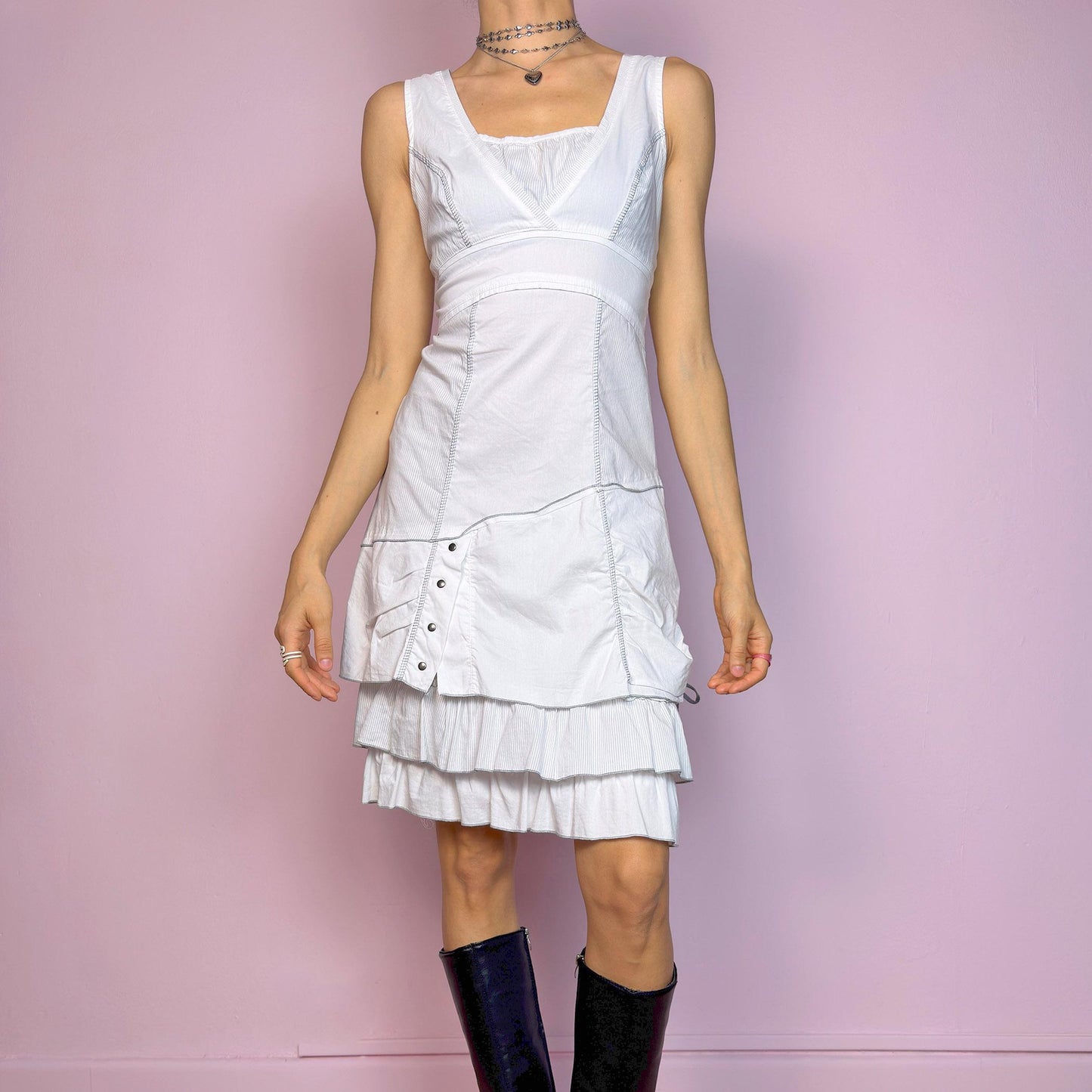 This vintage 2000s white sleeveless dress features a square neckline and sculpted panel seams. Made from a stretchy fabric, it includes a side zipper and falls to a midi length. Grommet hardware and a ruched drawstring side add a utilitarian twist, while the double-tiered ruffle hem softens the silhouette. Perfect for summer, this piece fuses deconstructed structure with a futuristic Y2K edge. Great vintage condition.