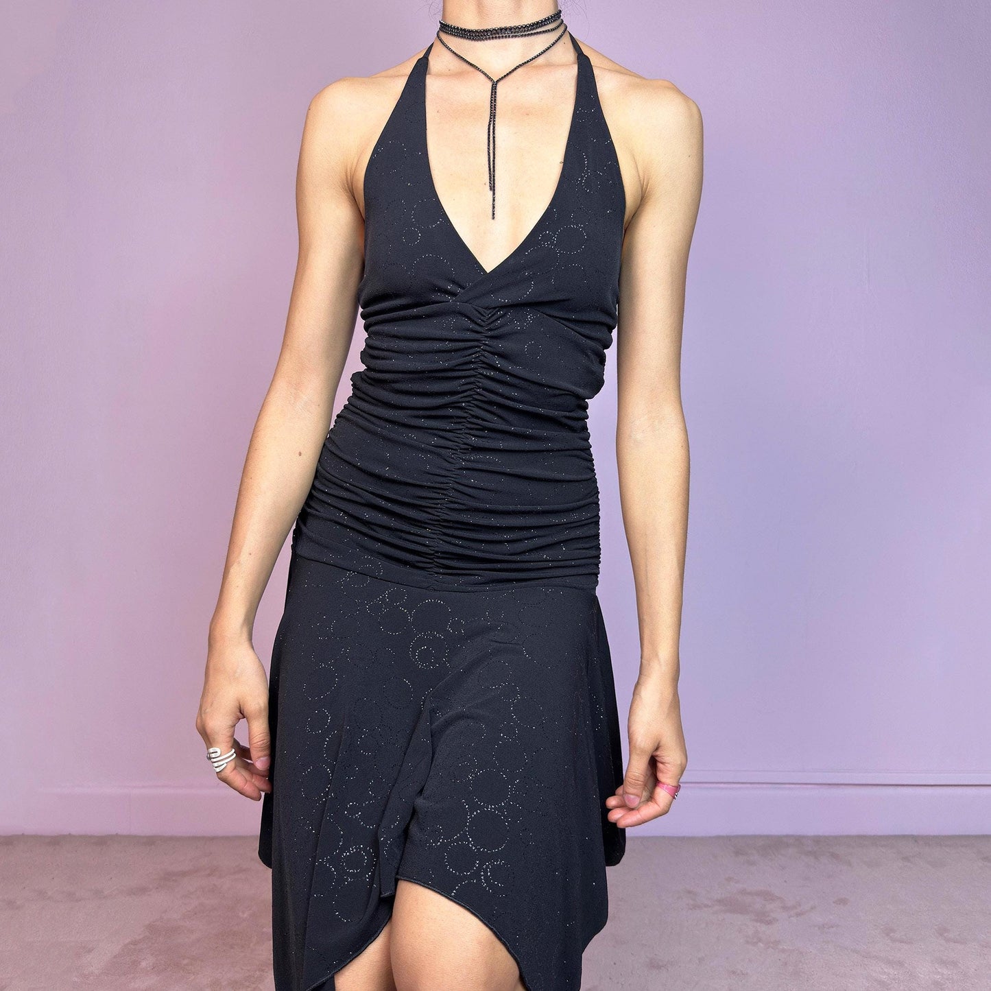 This vintage 2000s black dress features a halter tie neck, deep V-neckline, and allover subtle glitter swirl pattern. The ruched bodice sculpts the waist while the handkerchief hem adds movement and edge. Cut from stretchy fabric with a fluid, body-hugging drape. No closures. A cyber-glam silhouette with clubwear energy—perfect for parties, raves, or statement summer nights. Great vintage condition.
