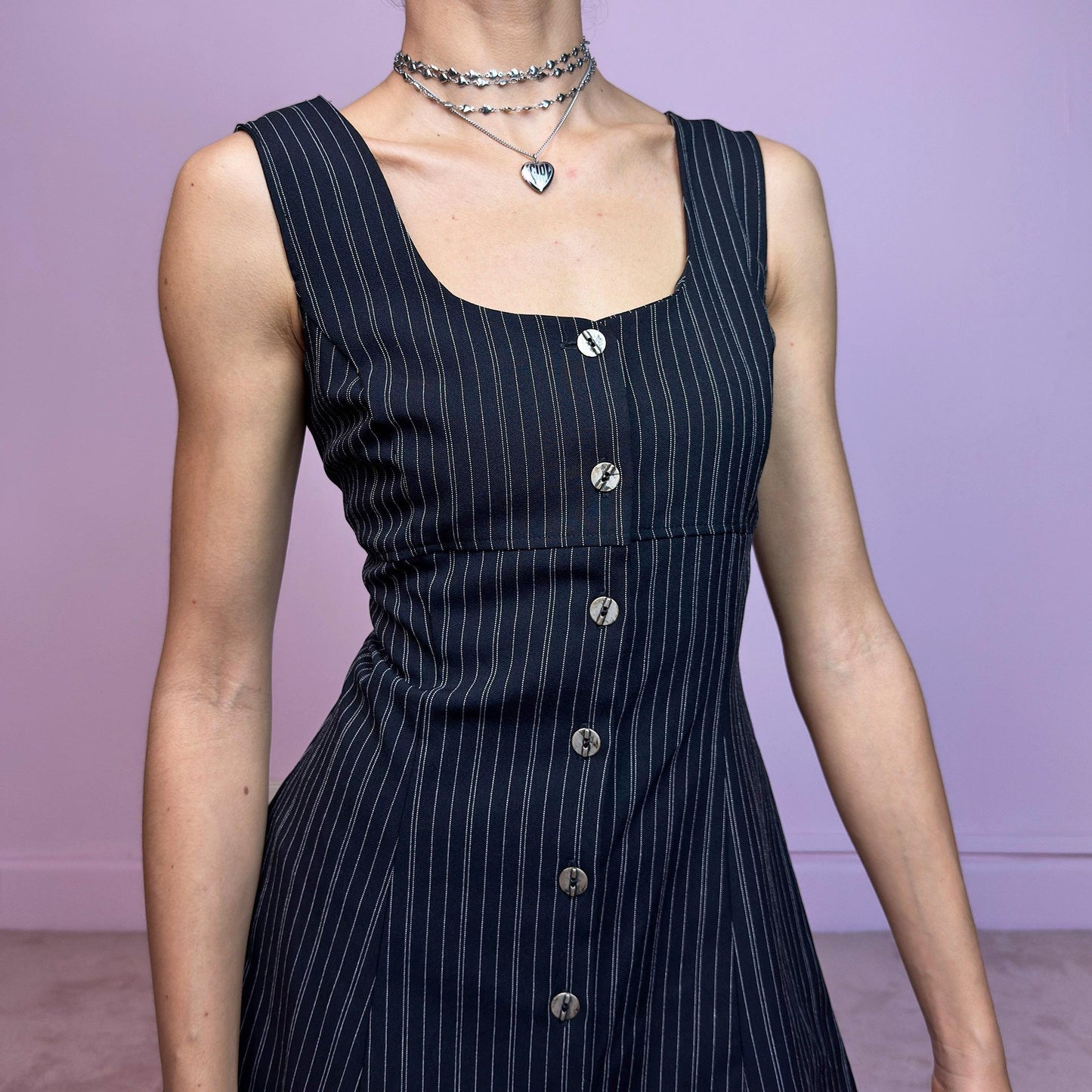 The Vintage 90s Black Pinstripe Mini Dress is a timeless piece with a flirty twist. This sleeveless dress features a black base with fine pinstripes, a fitted bodice with a square neckline, and a full front button closure. The slight A-line silhouette flatters the waist while keeping the vibe playful and polished. Ideal for 90s office-core and preppy looks. Excellent vintage condition.