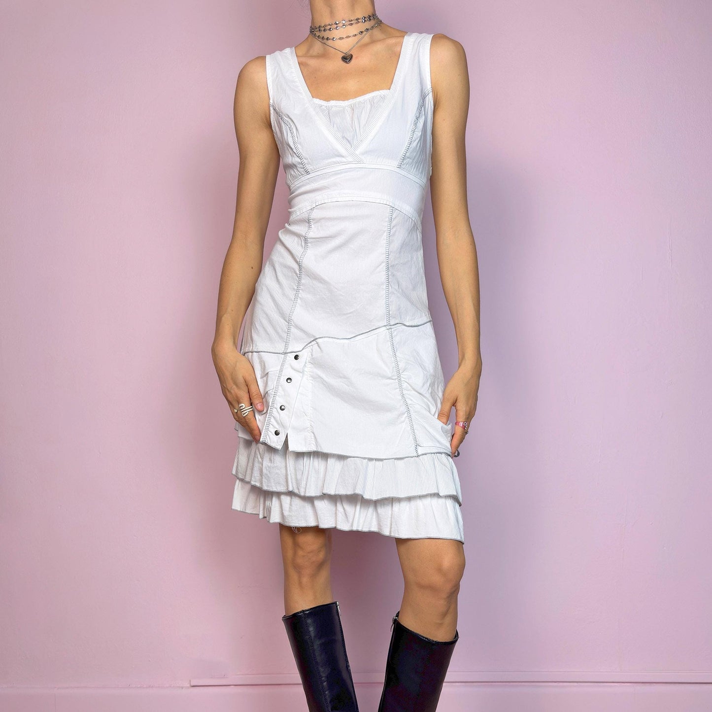 This vintage 2000s white sleeveless dress features a square neckline and sculpted panel seams. Made from a stretchy fabric, it includes a side zipper and falls to a midi length. Grommet hardware and a ruched drawstring side add a utilitarian twist, while the double-tiered ruffle hem softens the silhouette. Perfect for summer, this piece fuses deconstructed structure with a futuristic Y2K edge. Great vintage condition.