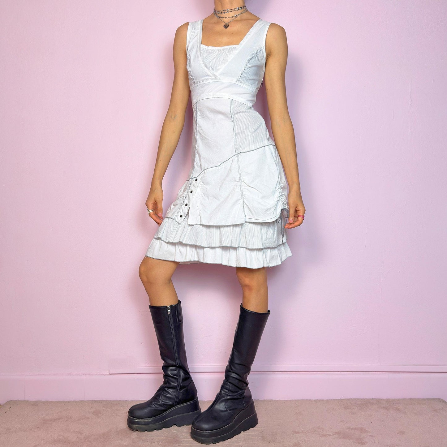 This vintage 2000s white sleeveless dress features a square neckline and sculpted panel seams. Made from a stretchy fabric, it includes a side zipper and falls to a midi length. Grommet hardware and a ruched drawstring side add a utilitarian twist, while the double-tiered ruffle hem softens the silhouette. Perfect for summer, this piece fuses deconstructed structure with a futuristic Y2K edge. Great vintage condition.