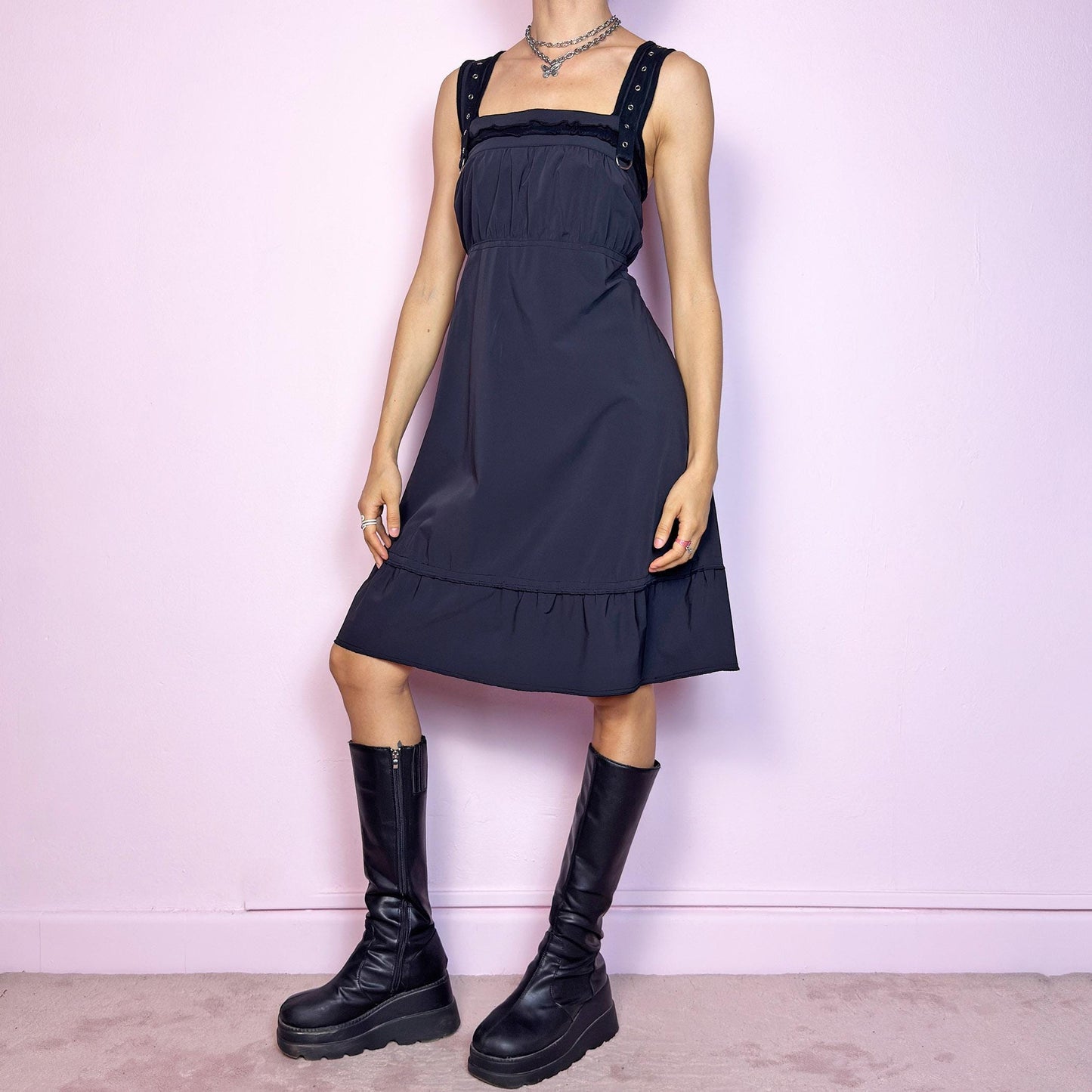This vintage 2000s black sleeveless dress features a gathered bust, square neckline, wide grommet straps with silver hardware and a back zipper closure. The structured fabric has slight stretch, while the A-line silhouette falls to midi length with a subtle ruffle hem. A utilitarian-meets-feminine Y2K piece—minimalist and wearable with a cyber futuristic edge. Perfect for everyday summer styling with impact.