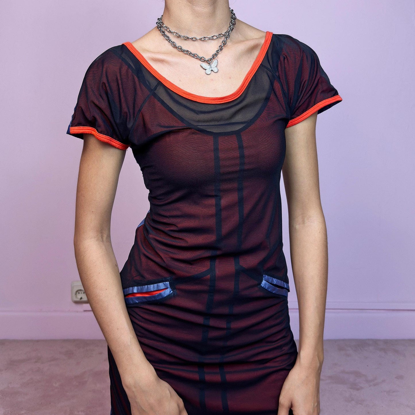 This vintage 2000s mini dress features sheer black mesh over an orange base with blue vertical stripes. Glossy faux-pocket details and neon orange trim add bold contrast. The fitted silhouette includes short sleeves, a scoop neckline, and stretch throughout. A cyber-inspired Y2K piece with futuristic, sporty edge—perfect for summer nights and bold party looks.