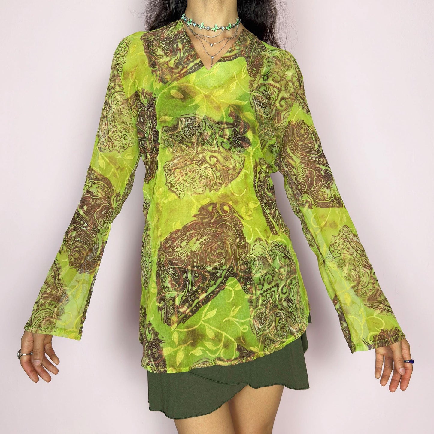 This vintage 2000s bohemian sheer green blouse casts a psychedelic glow with swirling brown paisley motifs and fluid draping. Flared long sleeves and a soft V-neckline evoke carefree elegance. Wear it layered at summer parties or as a lightweight, kaftan-inspired beach cover-up. Made in France from lightweight polyester. Excellent vintage condition.