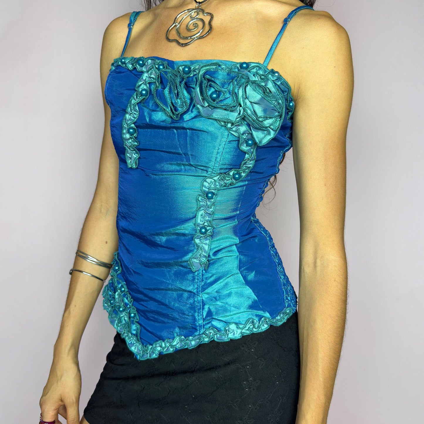 This vintage 2000s iridescent turquoise corset top features adjustable spaghetti straps, floral rosette appliqué, and beaded trim. The structured front sculpts the waist, while the ruched smocked back adds stretch and comfort. An asymmetrical hem and glossy finish elevate it for prom, weddings, or dreamy nights out. A romantic Y2K formal top that blends fairytale opulence with a bold, sculptural silhouette. Excellent vintage condition.