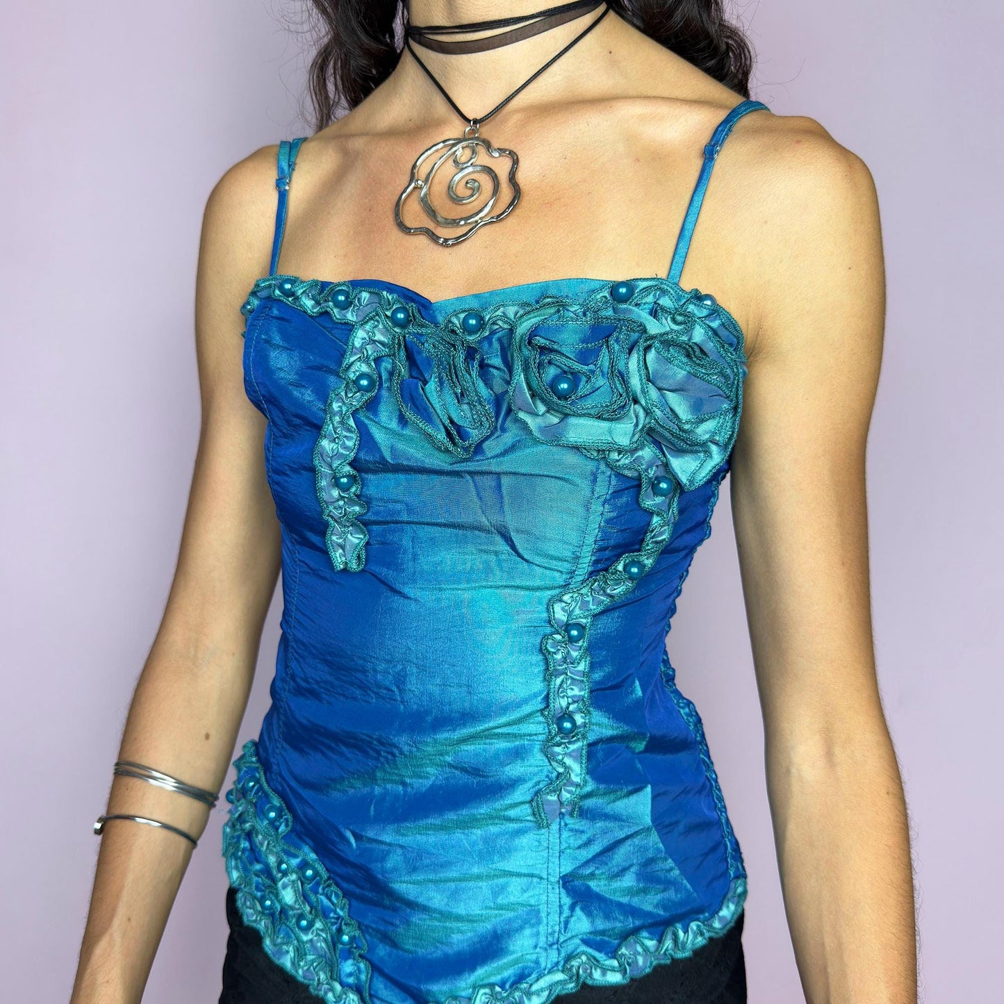 This vintage 2000s iridescent turquoise corset top features adjustable spaghetti straps, floral rosette appliqué, and beaded trim. The structured front sculpts the waist, while the ruched smocked back adds stretch and comfort. An asymmetrical hem and glossy finish elevate it for prom, weddings, or dreamy nights out. A romantic Y2K formal top that blends fairytale opulence with a bold, sculptural silhouette. Excellent vintage condition.