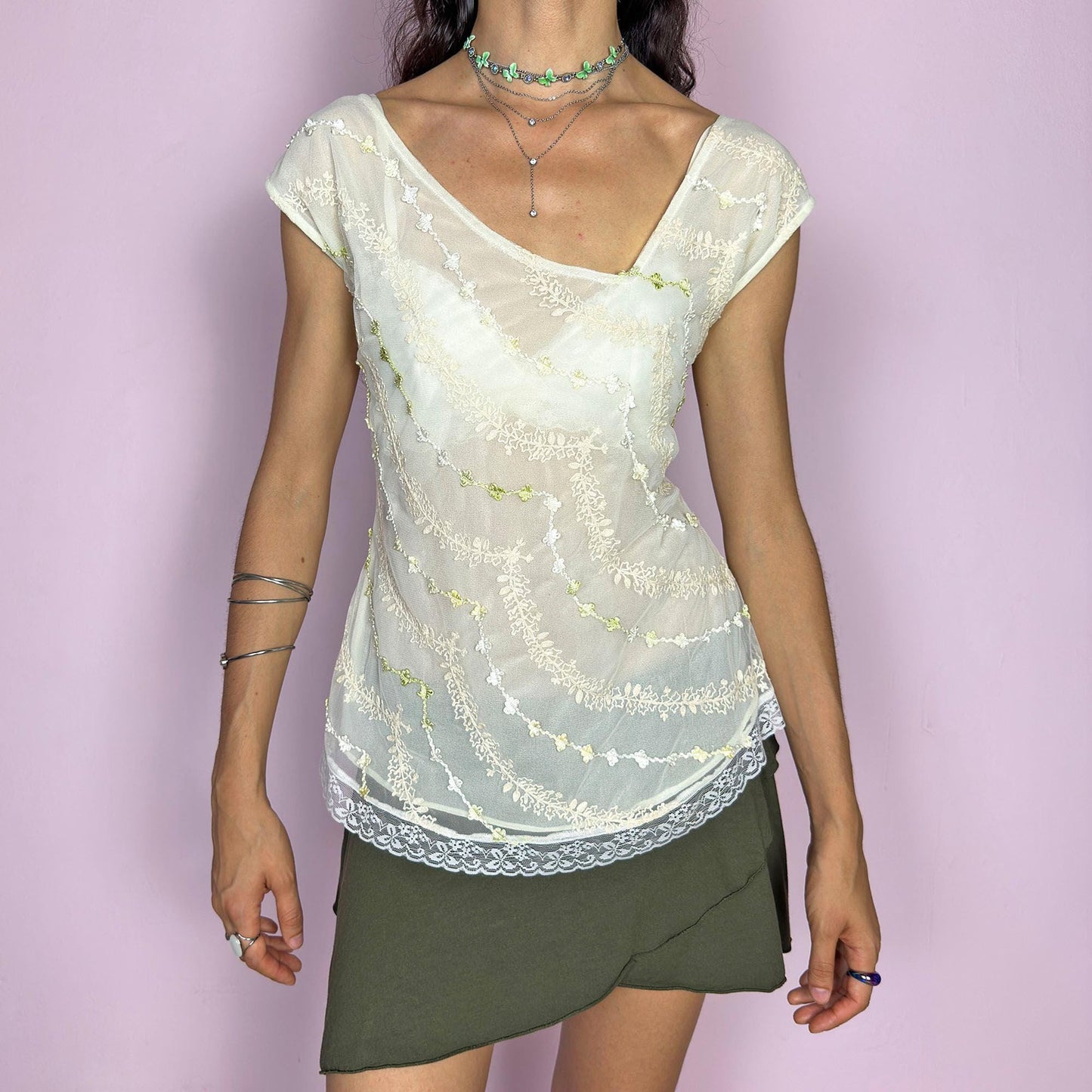 This vintage 2000s cream white mesh top features semi-sheer fabric with delicate embroidery in soft yellow, white, and pale green. The asymmetrical neckline and cap sleeves create an ethereal drape, while lace trim at the hem adds a romantic finish. This fairy-like Y2K blouse evokes lightness and whimsy—perfect for dreamy summer looks, warm days, and delicate layering. Made of polyester blend.