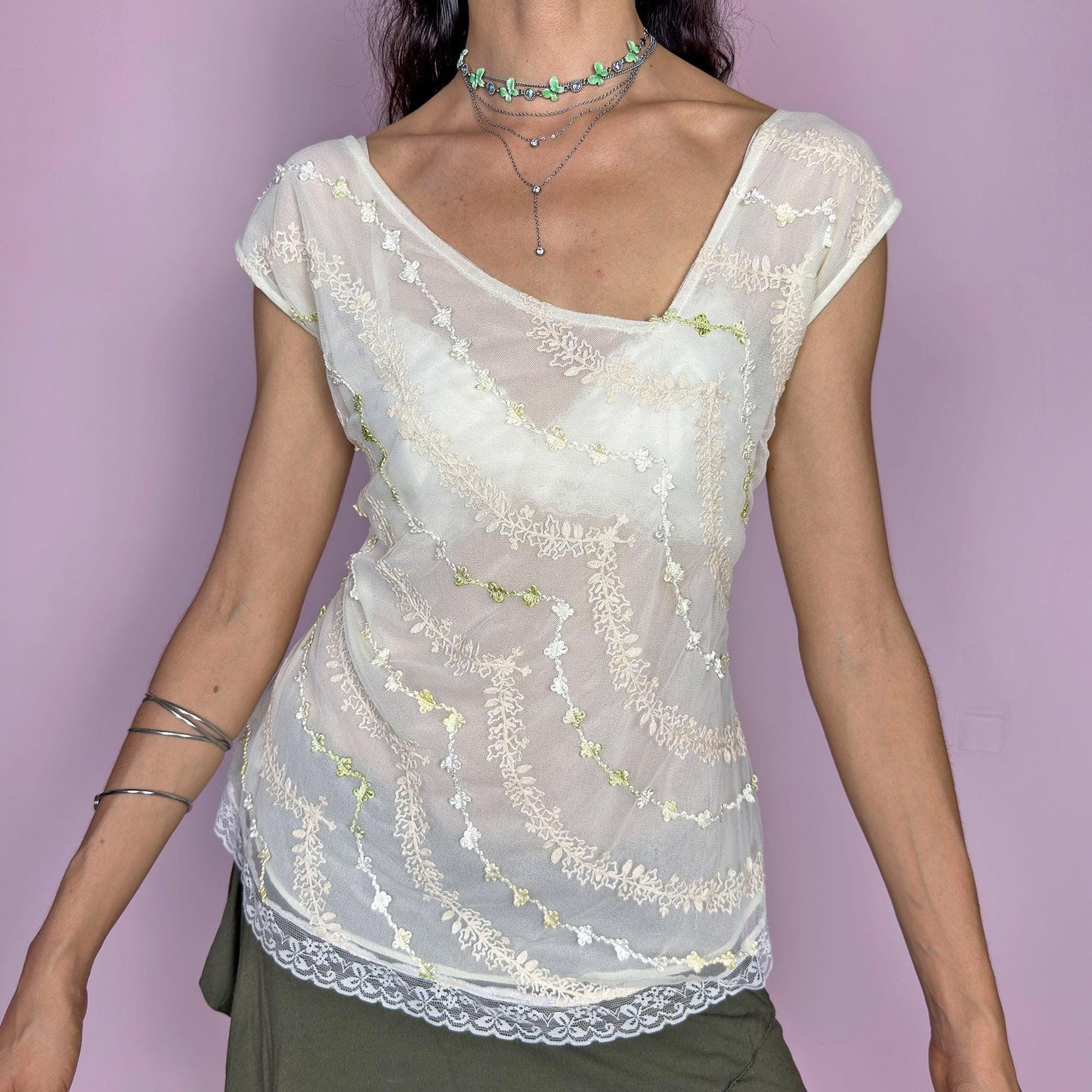 This vintage 2000s cream white mesh top features semi-sheer fabric with delicate embroidery in soft yellow, white, and pale green. The asymmetrical neckline and cap sleeves create an ethereal drape, while lace trim at the hem adds a romantic finish. This fairy-like Y2K blouse evokes lightness and whimsy—perfect for dreamy summer looks, warm days, and delicate layering. Made of polyester blend.