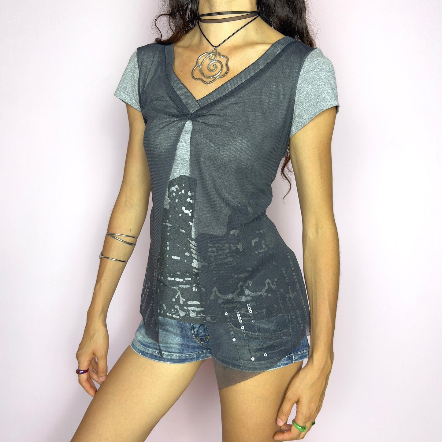 The Y2K Gray Cityscape Mesh Top is a vintage 2000s piece featuring a skyline motif and a semi-sheer mesh overlay with subtle sequin details. Designed with a V-neckline, short sleeves, and a soft front slit, it creates a deconstructed, layered silhouette. Minimal yet visually striking, it channels the energy of night cityscapes. Perfect for casual summer styling or off-duty urban looks. Great vintage condition.