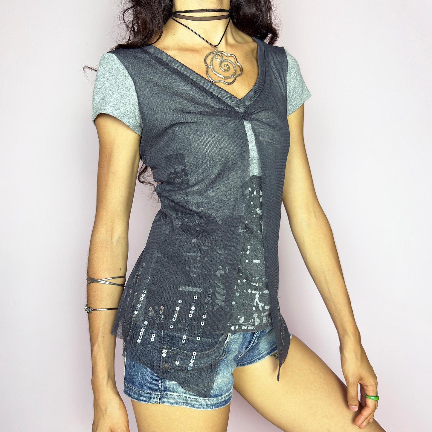 The Y2K Gray Cityscape Mesh Top is a vintage 2000s piece featuring a skyline motif and a semi-sheer mesh overlay with subtle sequin details. Designed with a V-neckline, short sleeves, and a soft front slit, it creates a deconstructed, layered silhouette. Minimal yet visually striking, it channels the energy of night cityscapes. Perfect for casual summer styling or off-duty urban looks. Great vintage condition.