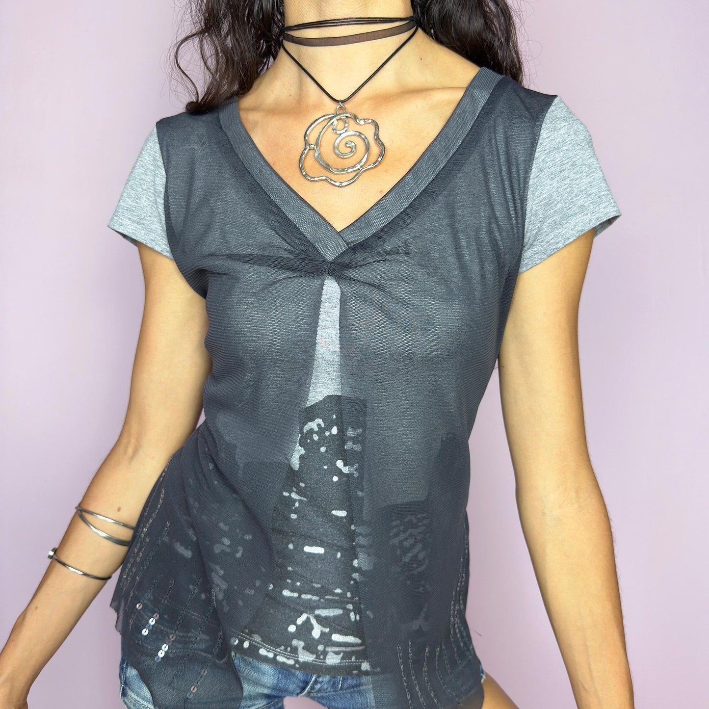 The Y2K Gray Cityscape Mesh Top is a vintage 2000s piece featuring a skyline motif and a semi-sheer mesh overlay with subtle sequin details. Designed with a V-neckline, short sleeves, and a soft front slit, it creates a deconstructed, layered silhouette. Minimal yet visually striking, it channels the energy of night cityscapes. Perfect for casual summer styling or off-duty urban looks. Great vintage condition.
