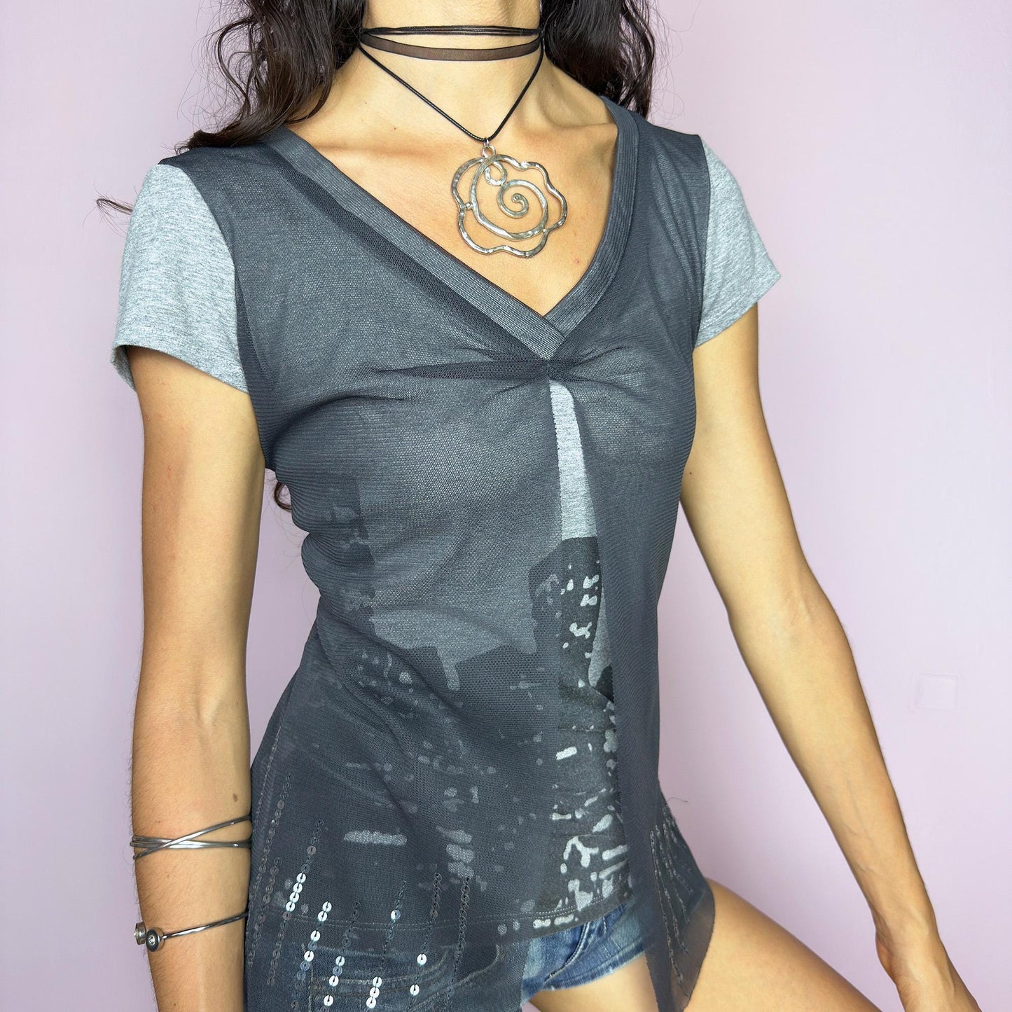 The Y2K Gray Cityscape Mesh Top is a vintage 2000s piece featuring a skyline motif and a semi-sheer mesh overlay with subtle sequin details. Designed with a V-neckline, short sleeves, and a soft front slit, it creates a deconstructed, layered silhouette. Minimal yet visually striking, it channels the energy of night cityscapes. Perfect for casual summer styling or off-duty urban looks. Great vintage condition.